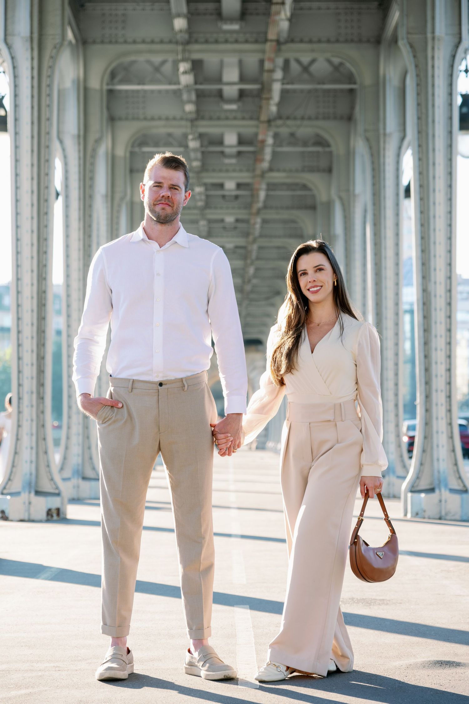 A stylish couple poses holding hands under an architectural bridge structure.