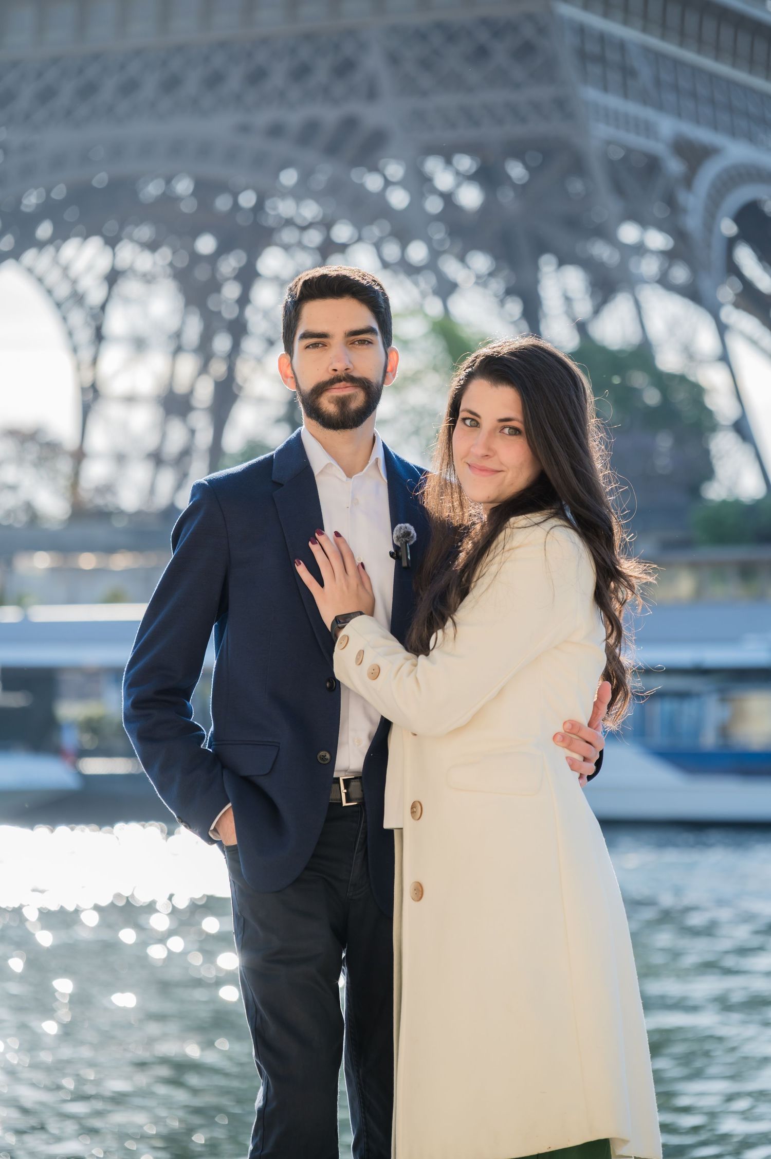 A couple poses romantically in front of the Eiffel Tower in Paris, wearing elegant navy suit and cream coat.