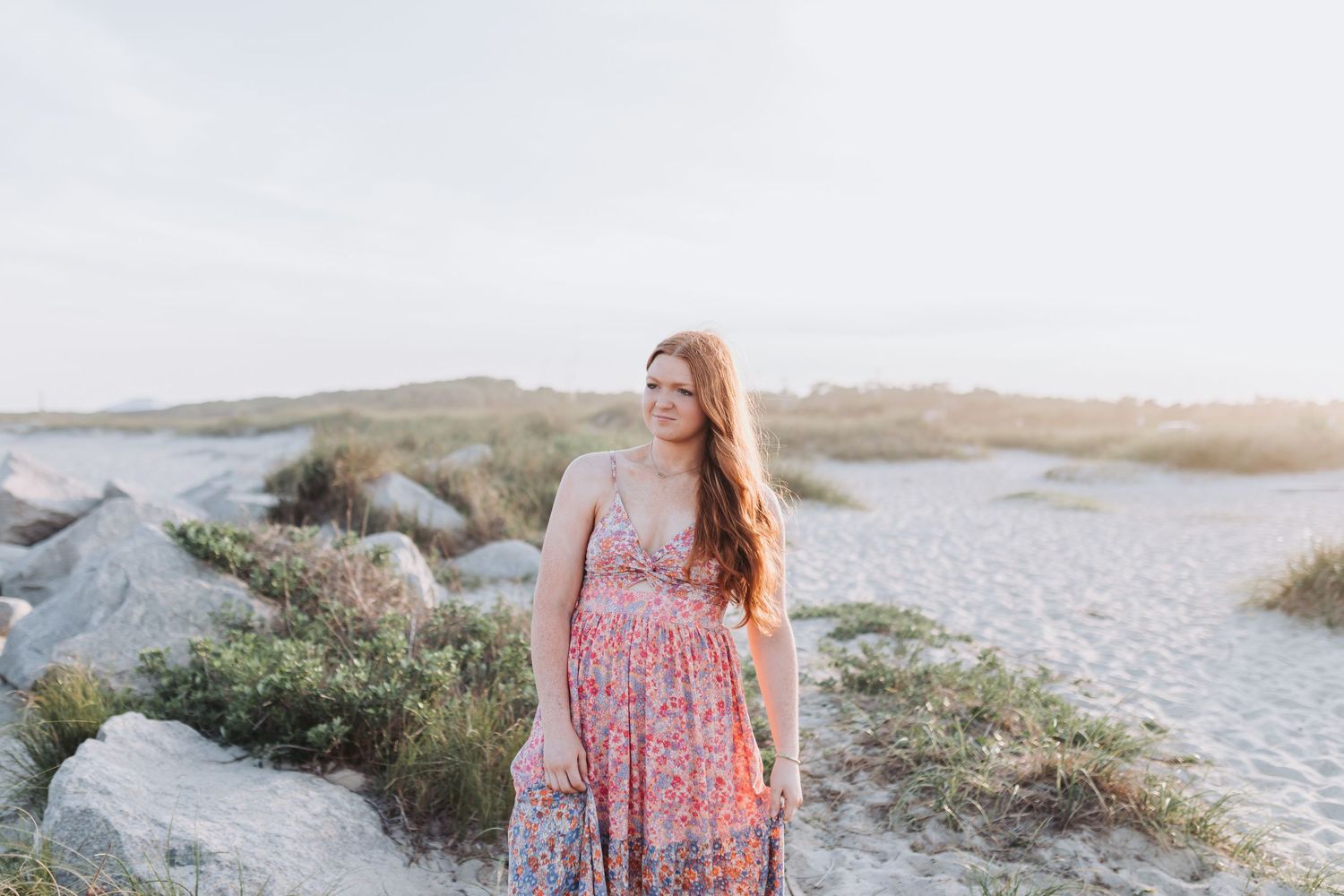 Someone in a floral maxi dress stands on a rocky coastal path during golden hour sunset with water visible in background.