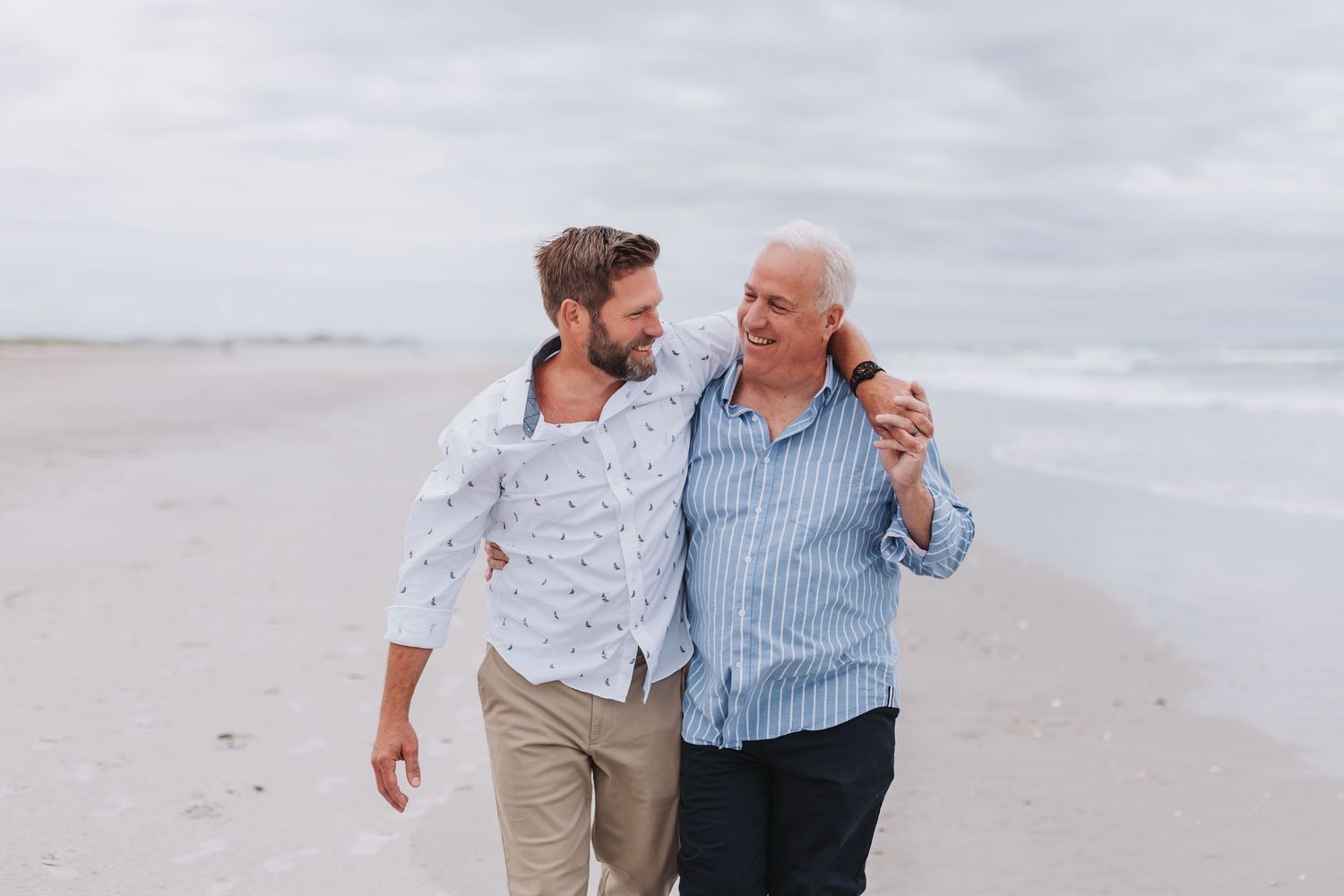 Two people walk together while smiling and embracing on a cloudy beach with soft sand and ocean waves.