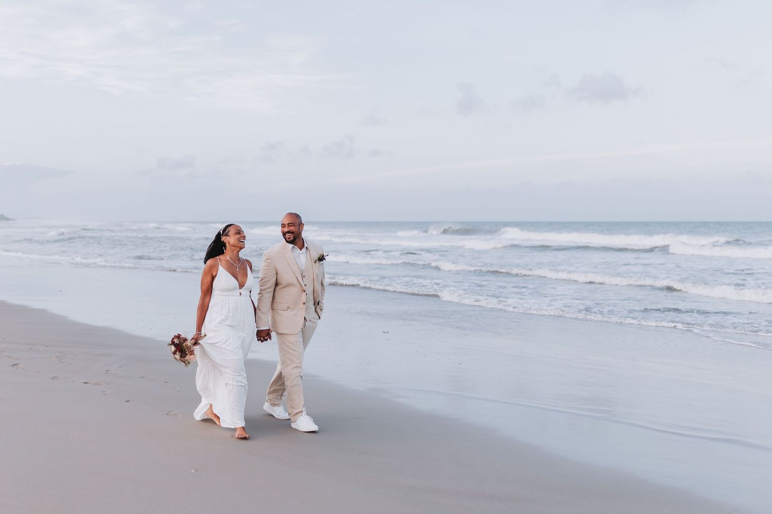 A couple in wedding attire walks together along a misty beach shoreline at sunset while waves roll in.