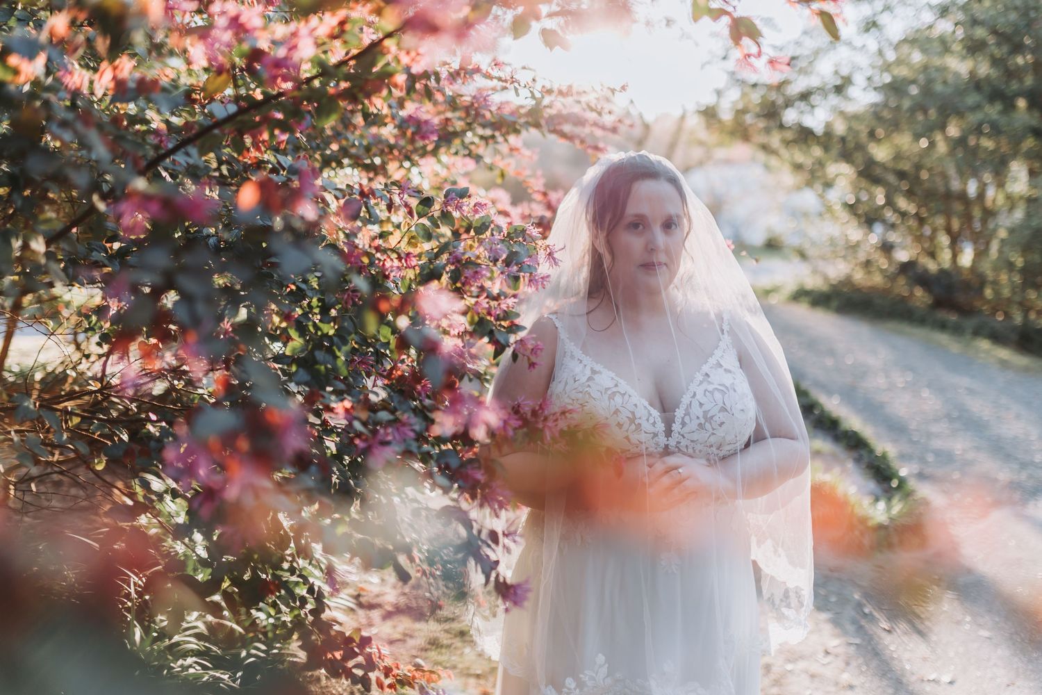 A dreamy bridal portrait under blooming pink flowers with sunlit bokeh effects in a garden setting.