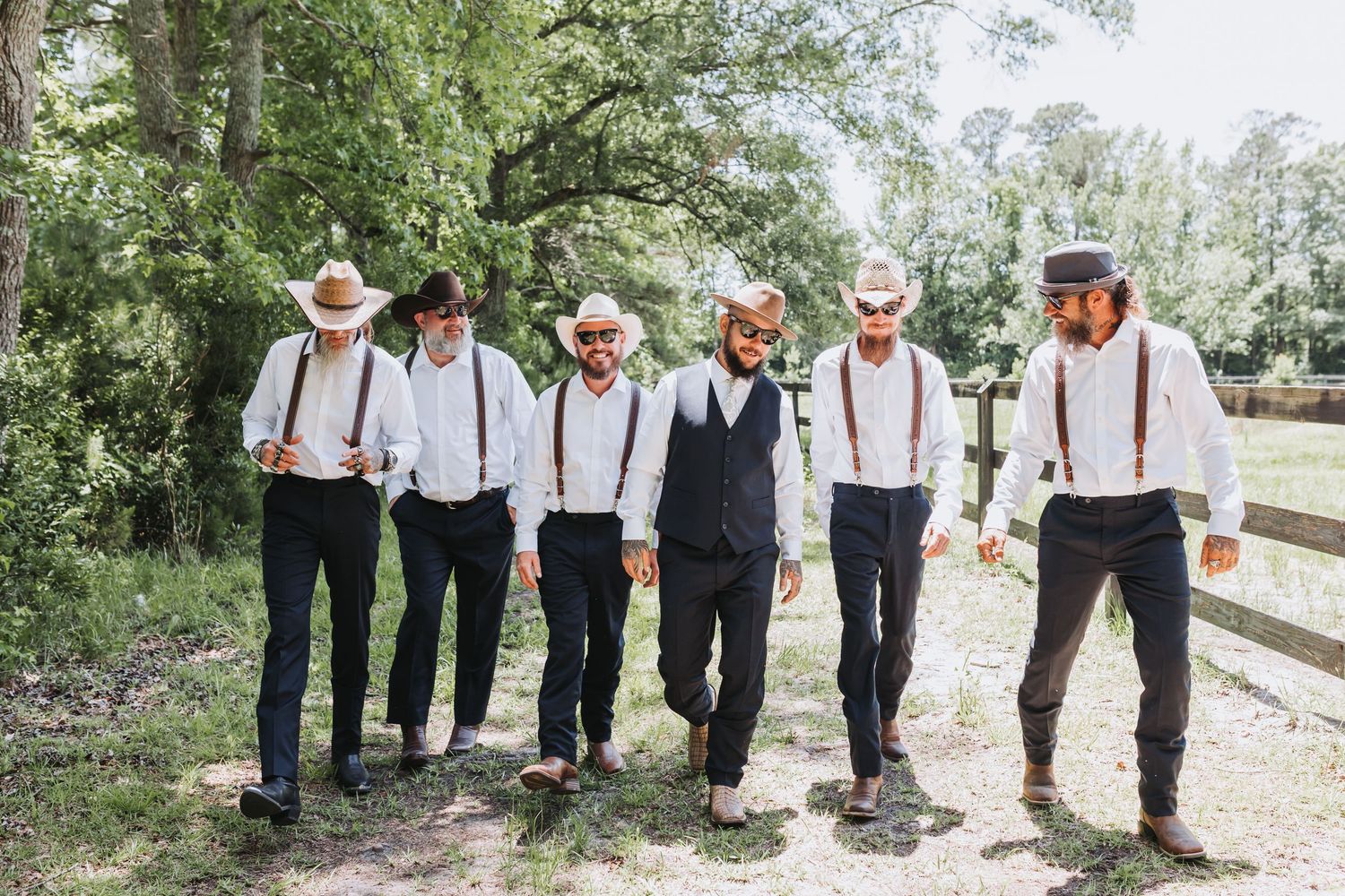 A group of groomsmen wearing suspenders, cowboy hats and boots pose together outdoors on a rustic path.