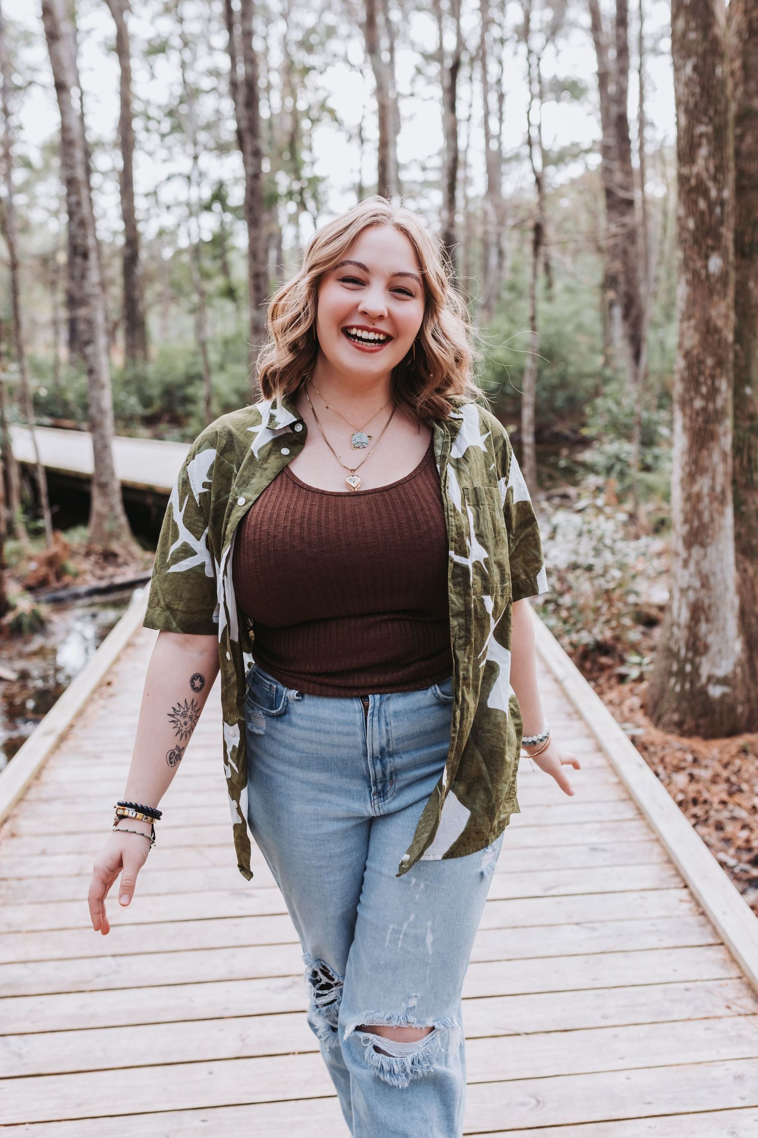 Person in casual outfit with camo jacket and ripped jeans standing on wooden boardwalk in forest setting.