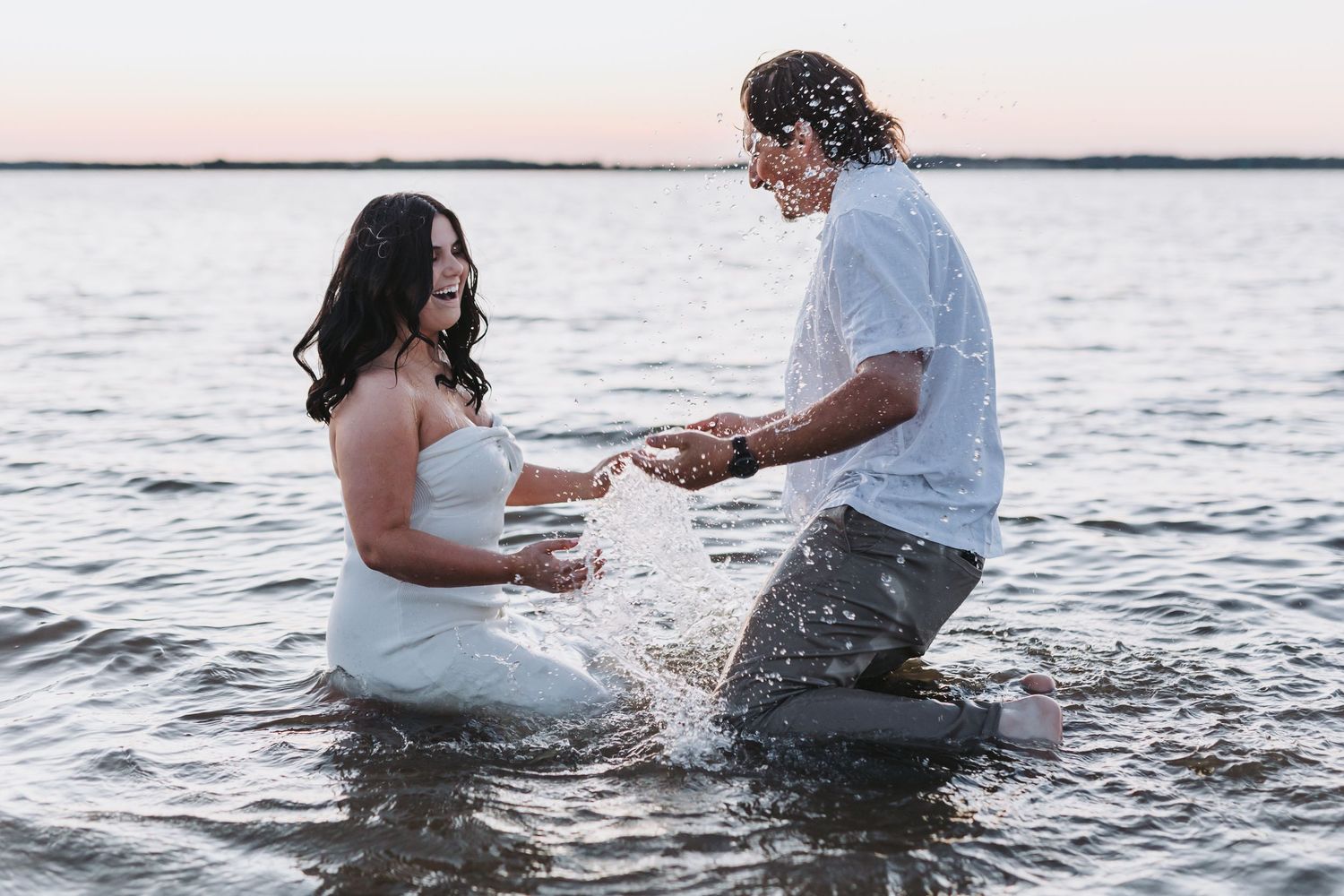 Two people in casual attire playfully splash in ocean water at sunset during a romantic photoshoot.