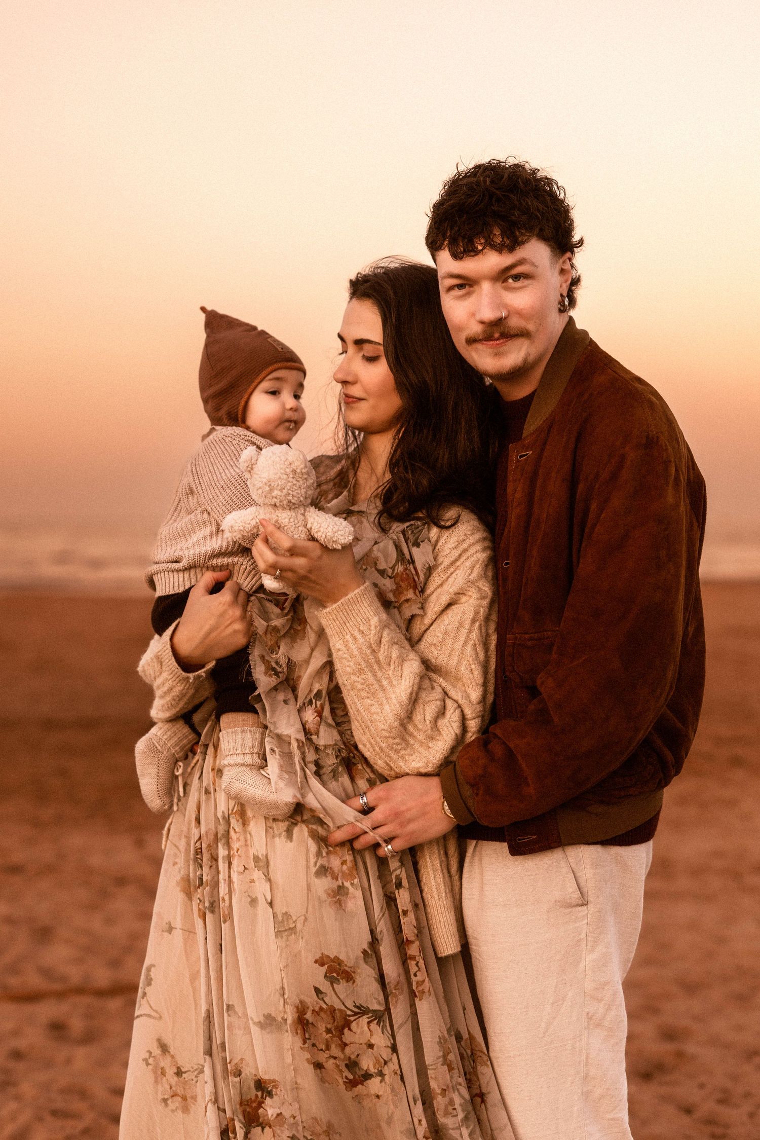 Family in warm earth tones stands together against sunset sky, baby wearing knit hat and parents in cozy sweaters. Javea photographer