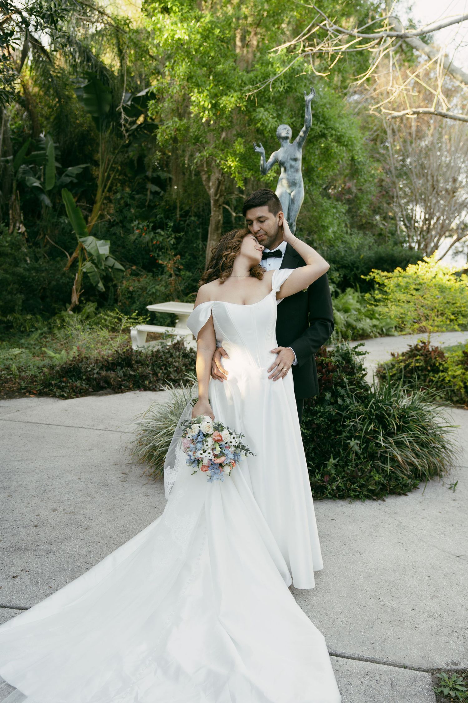 A newlywed couple poses in a garden with a statue, the bride holding a colorful bouquet.