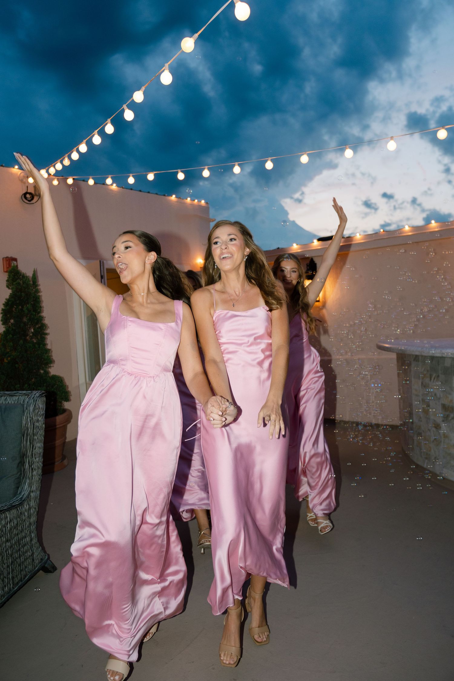 Bridesmaids in pink dresses dancing under string lights at dusk.