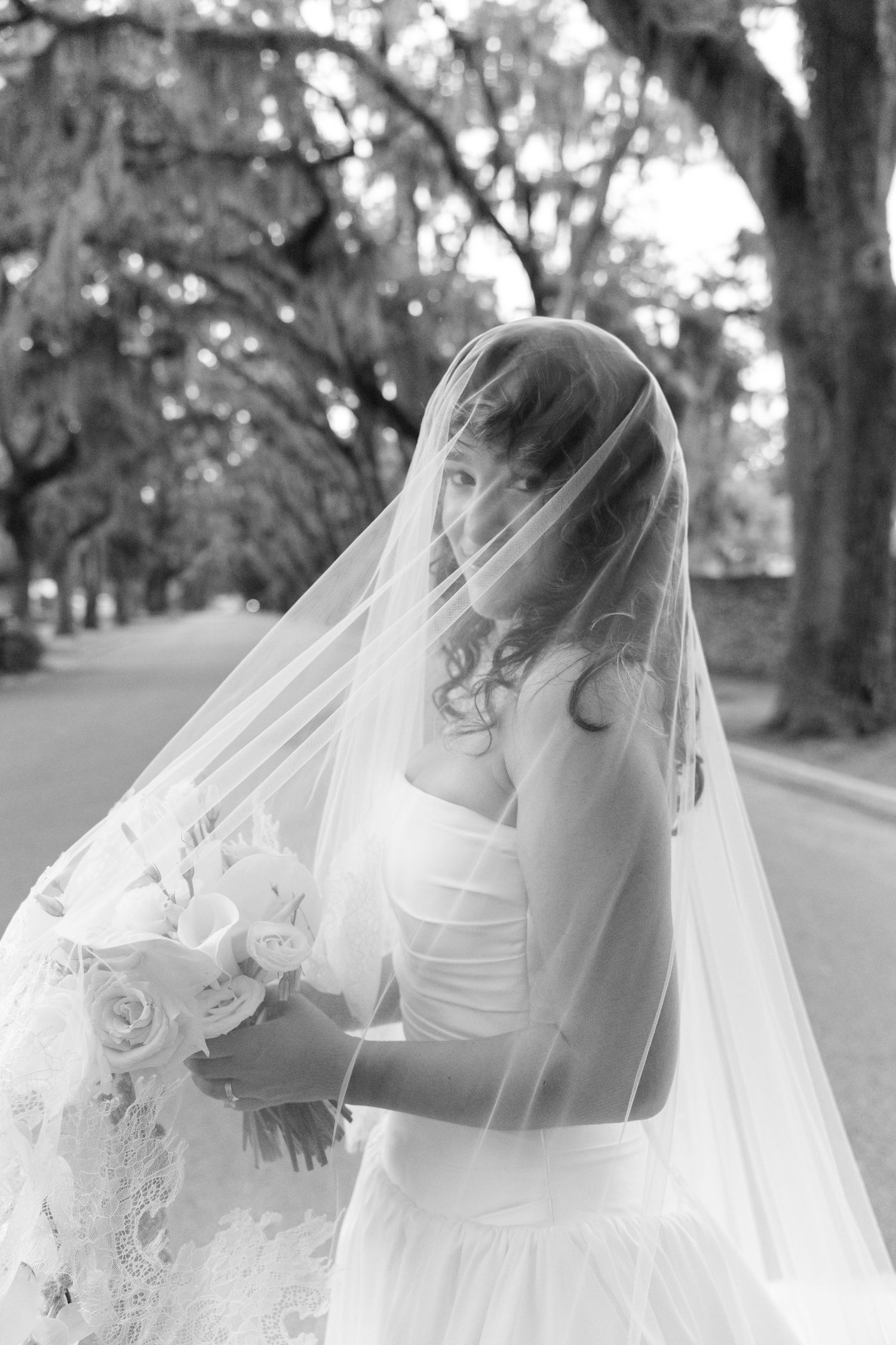 A bride in a white wedding dress and flowing veil poses on a tree-lined path in black and white photography.