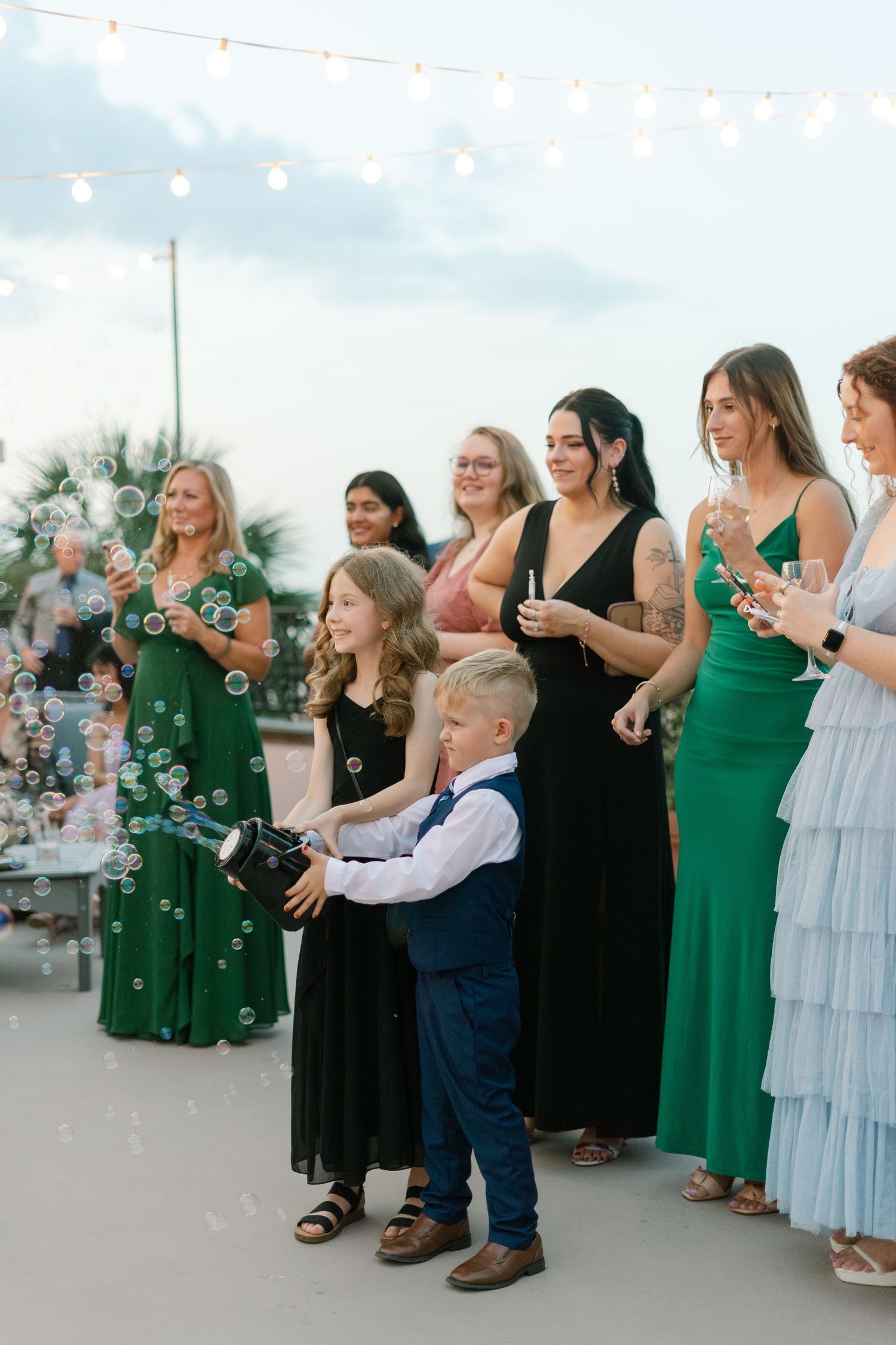 Wedding guests in formal attire blowing bubbles during an outdoor celebration.
