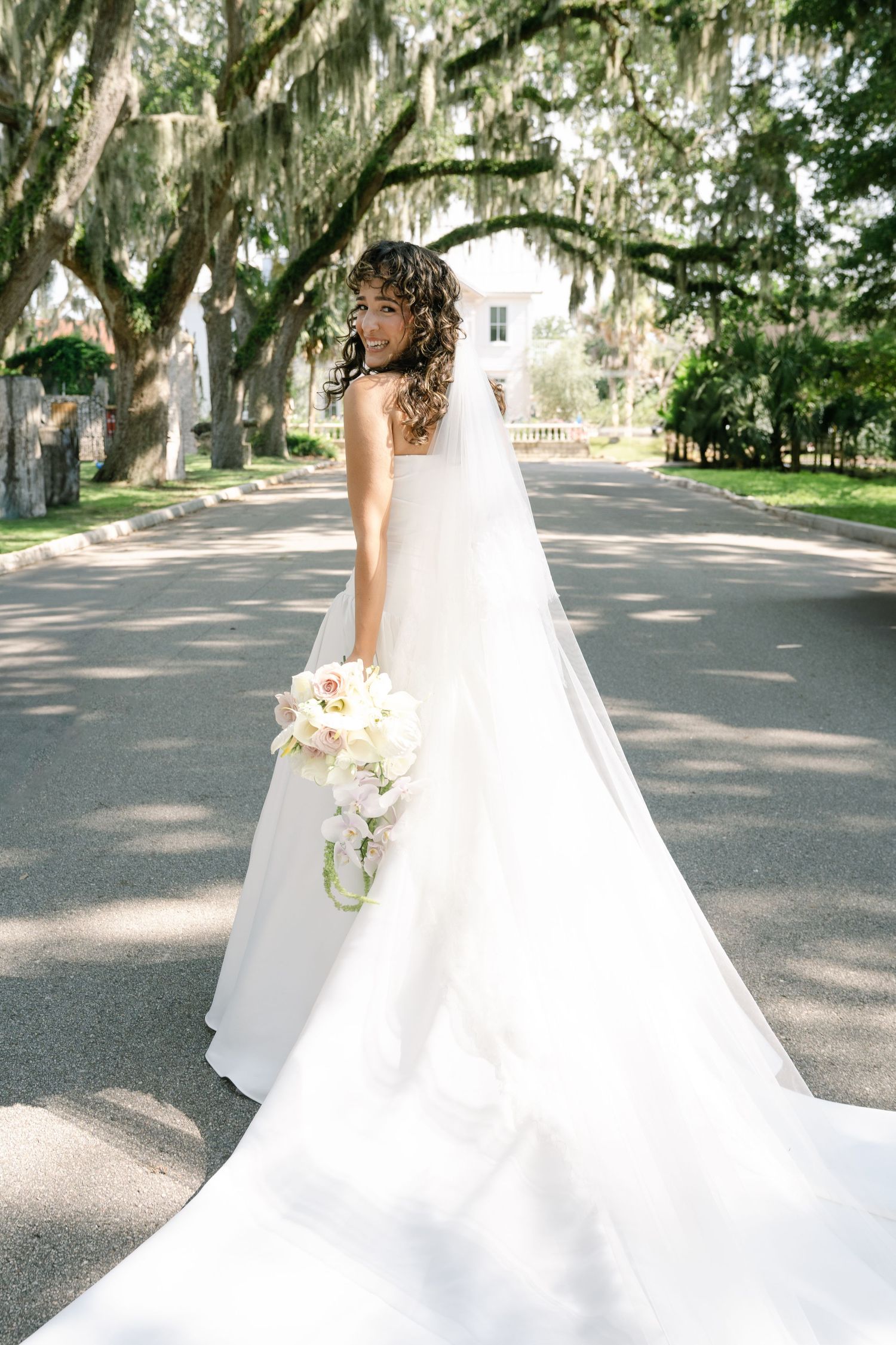 Bride in flowing white gown stands on a Spanish moss-lined avenue holding a delicate bouquet.