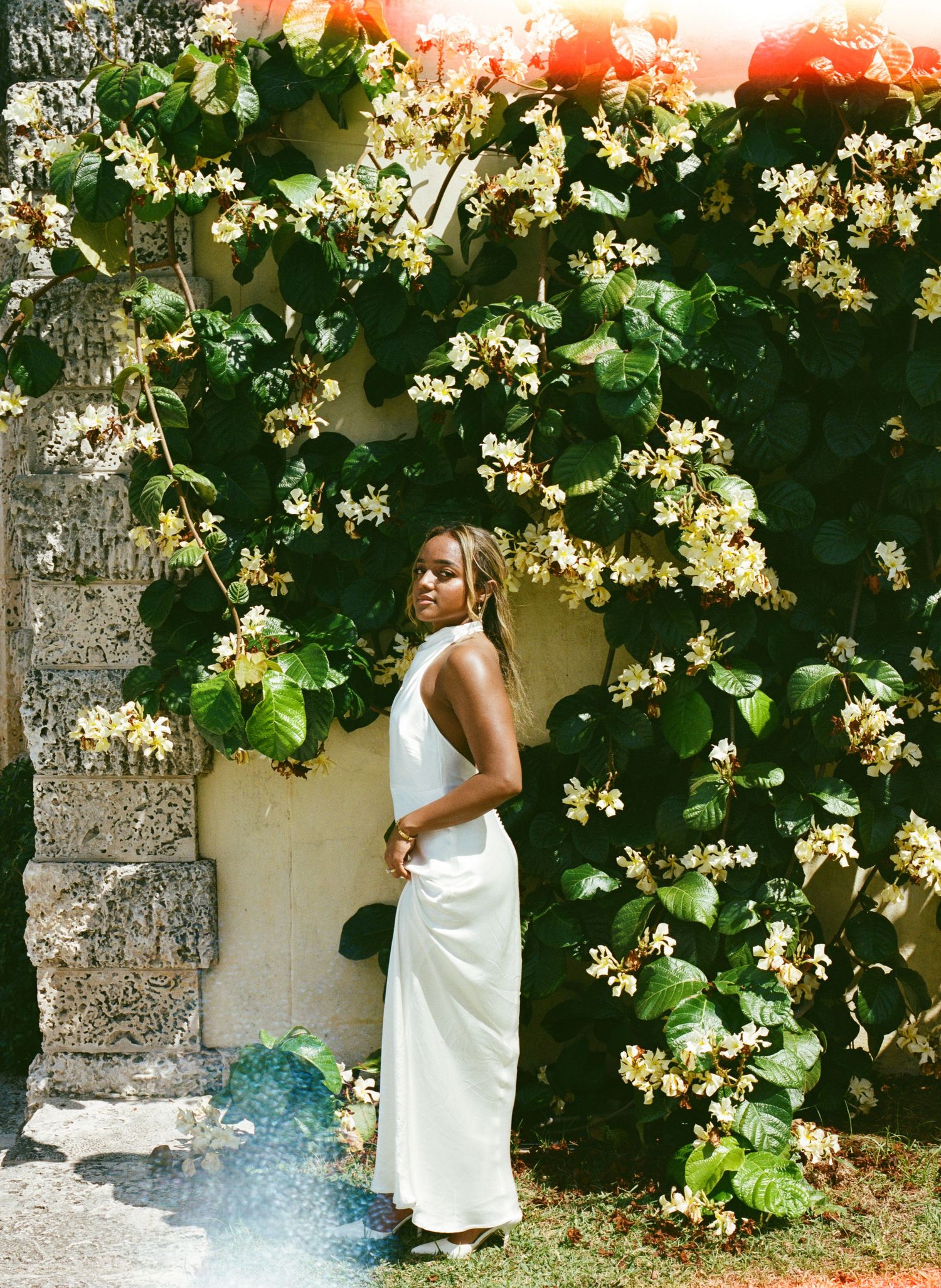 A figure in an elegant white dress poses against a wall covered in blooming white flowers and lush green vines.