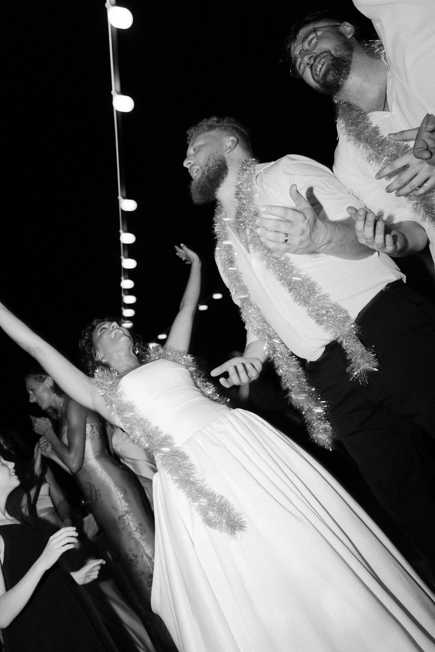 Black and white photo of energetic wedding dance moves under string lights.