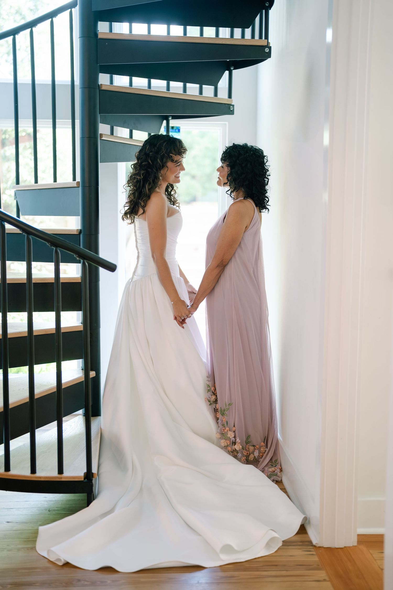 Two people in elegant gowns share an intimate moment on a modern spiral staircase.