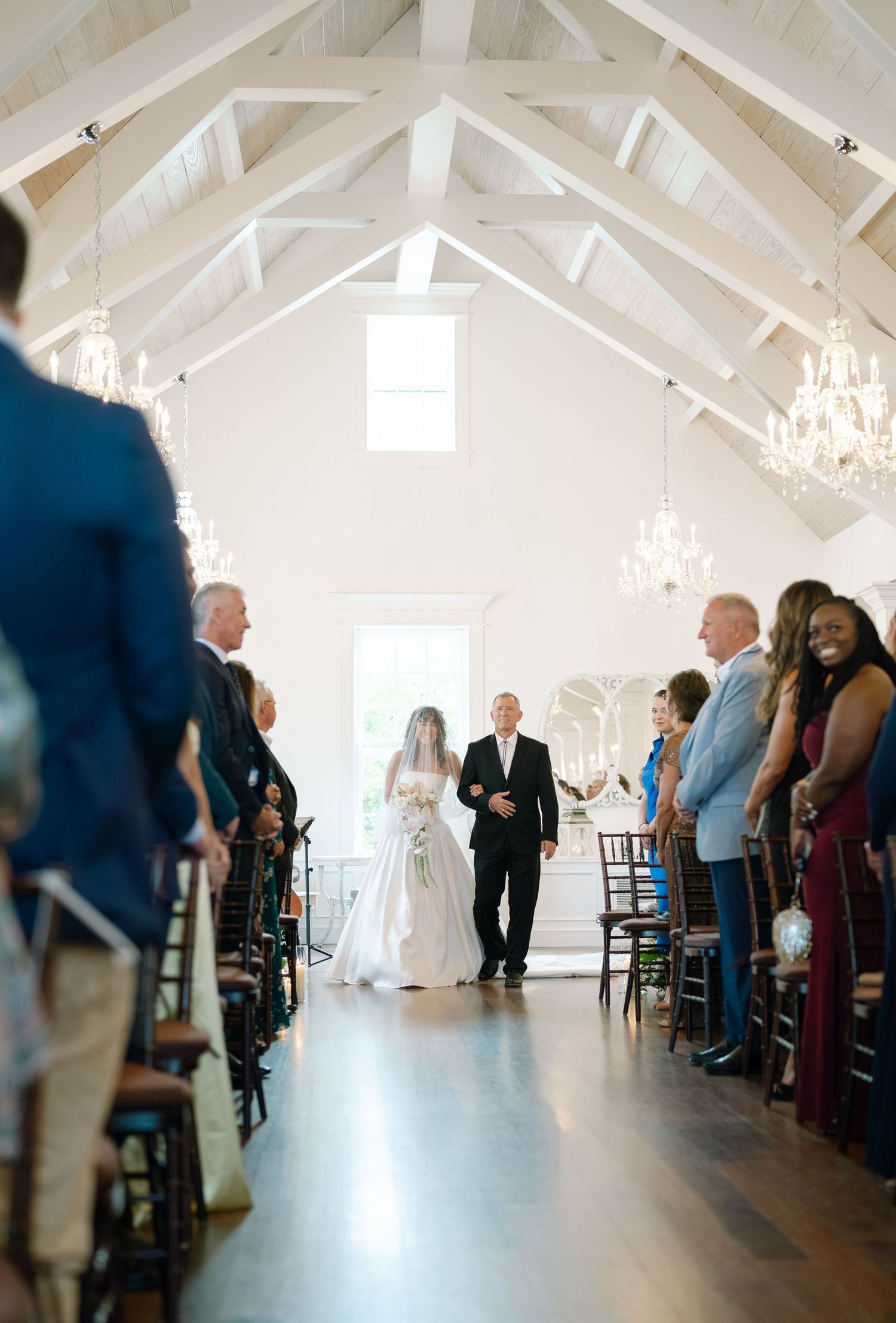 A wedding ceremony in a white vaulted chapel with crystal chandeliers and guests lining the aisle.