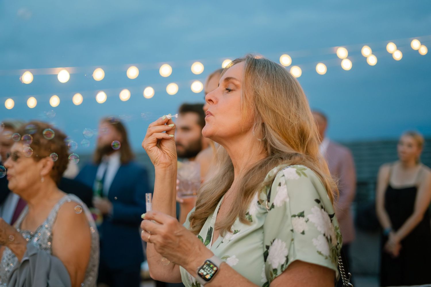 Guest blowing bubbles at twilight outdoor wedding reception under string lights.