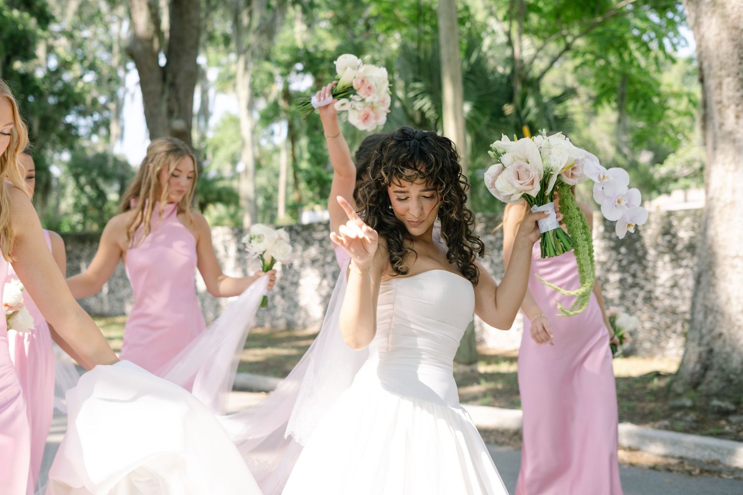 A bride in white celebrates with bridesmaids in pink dresses under moss-draped trees.
