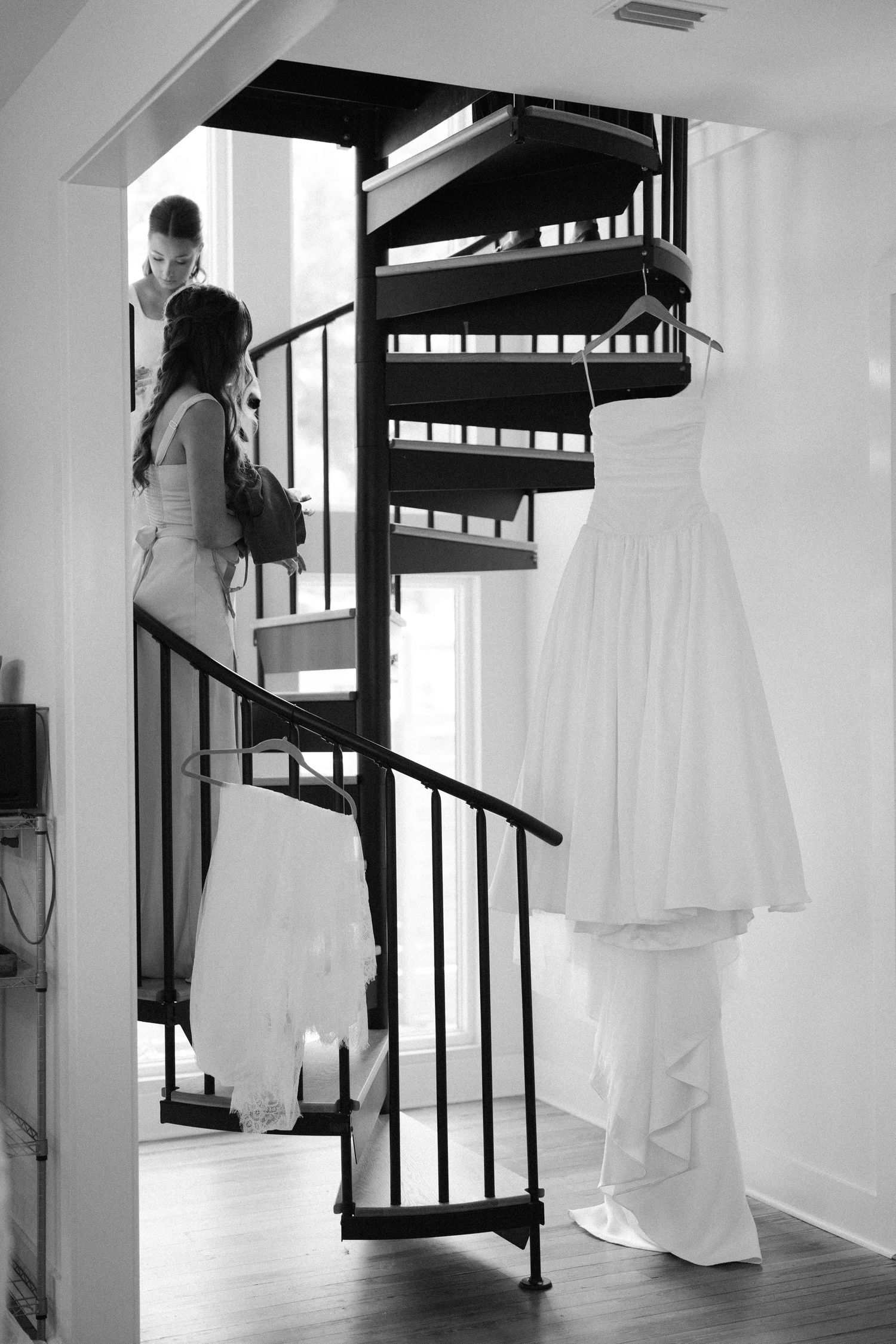 A wedding dress hangs elegantly beside a black spiral staircase in black and white photography.