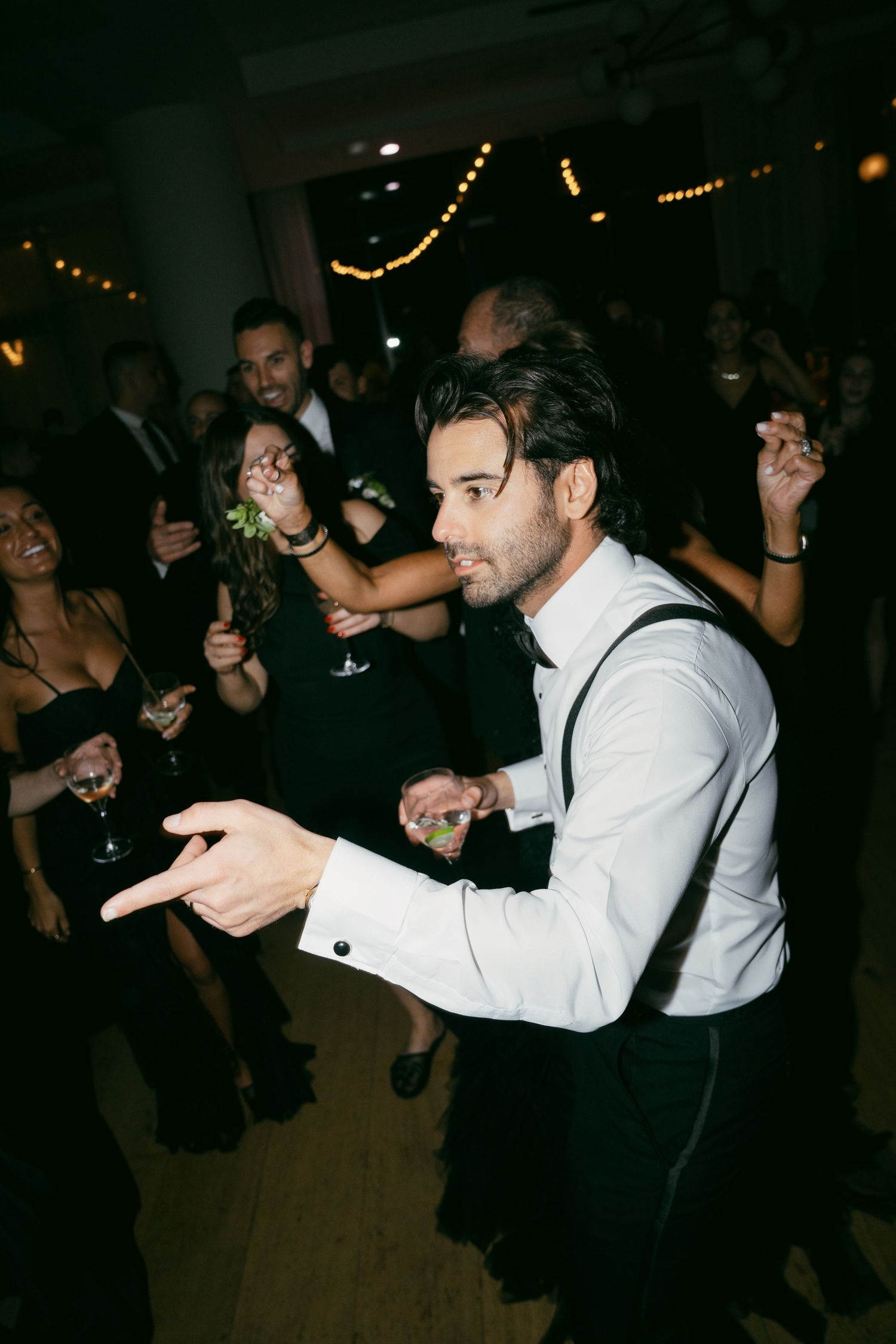 Partygoer in white shirt and suspenders dances energetically on dance floor with crowd in dark venue with string lights.