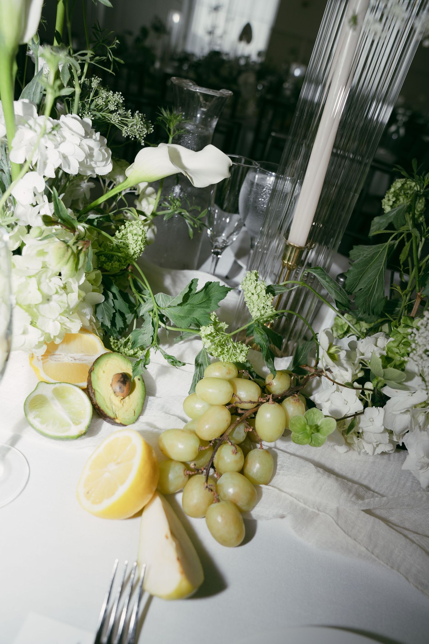 An elegant table setting with white flowers, grapes, citrus fruits, and avocado against a bright white tablecloth.
