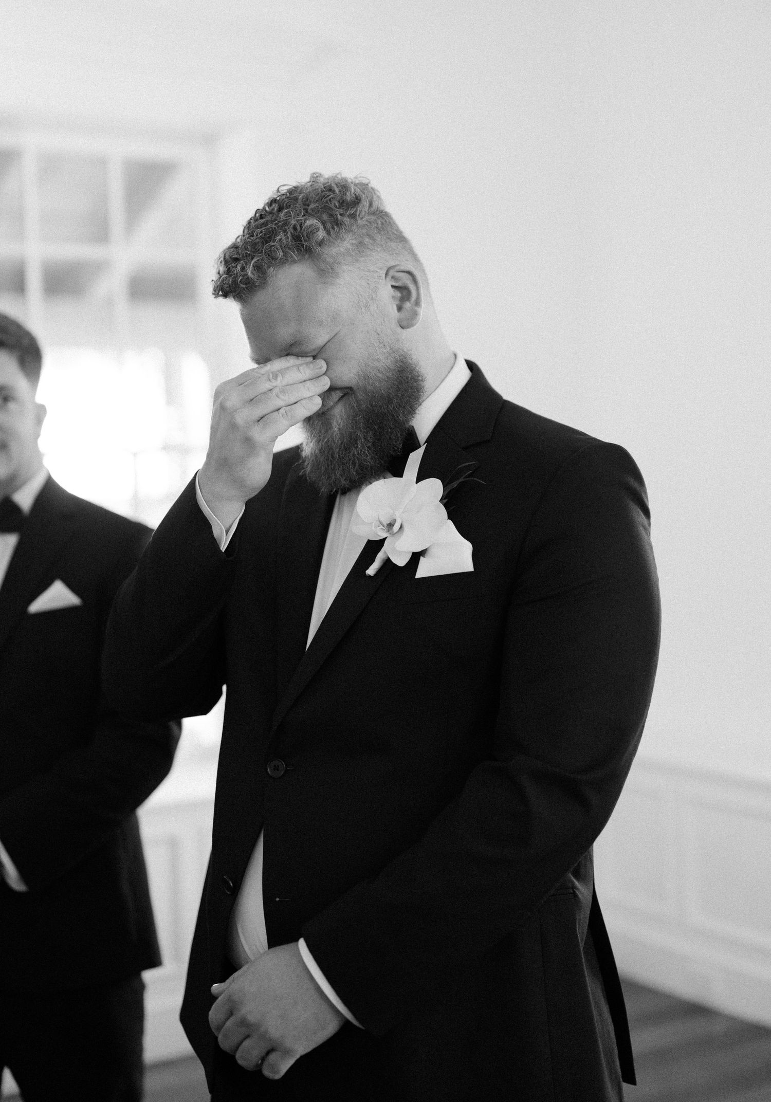 A bearded groom in a black tuxedo covers his face in an emotional moment during a wedding ceremony.