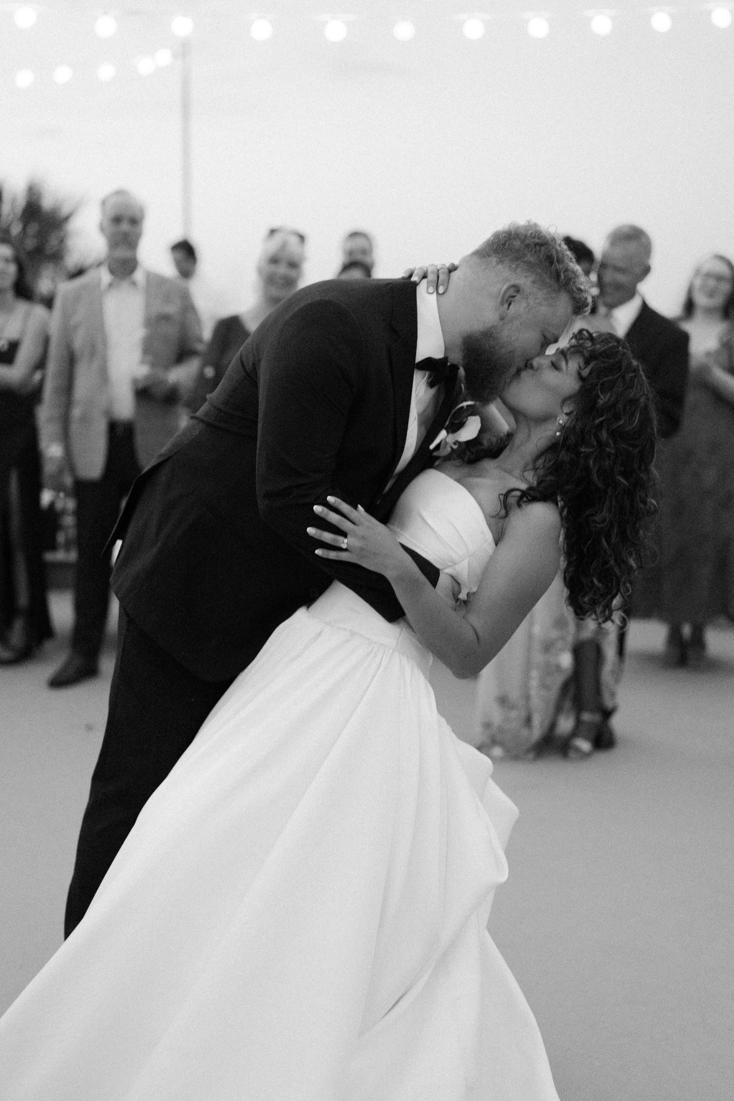 A romantic dip kiss between newlyweds during their wedding celebration with guests in the background.