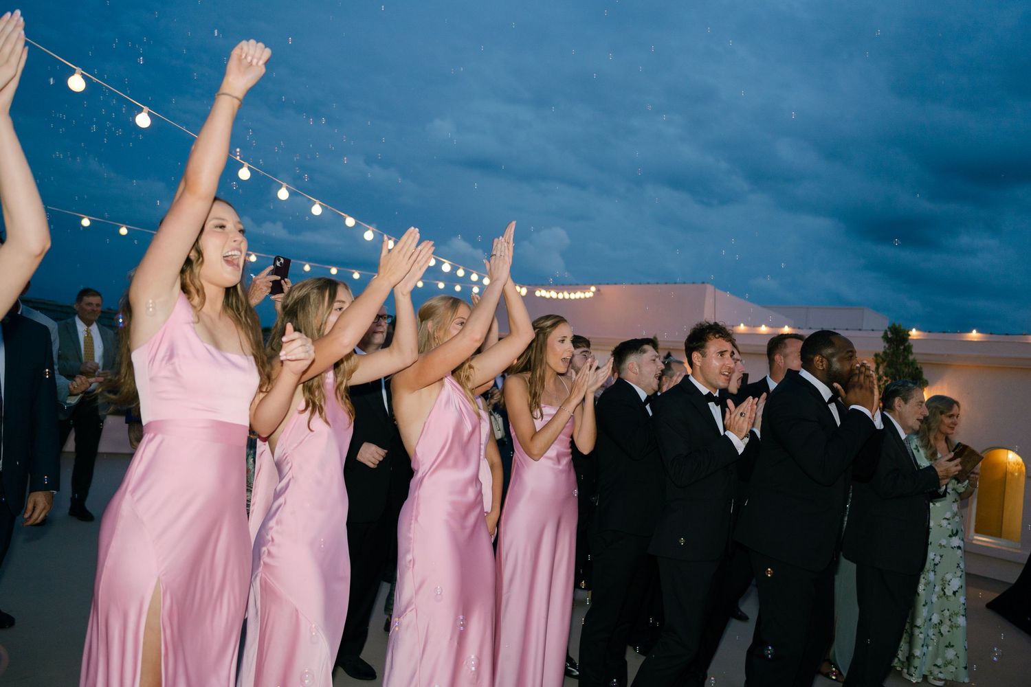 Bridesmaids in pink dresses and groomsmen celebrate with raised arms under evening sky.
