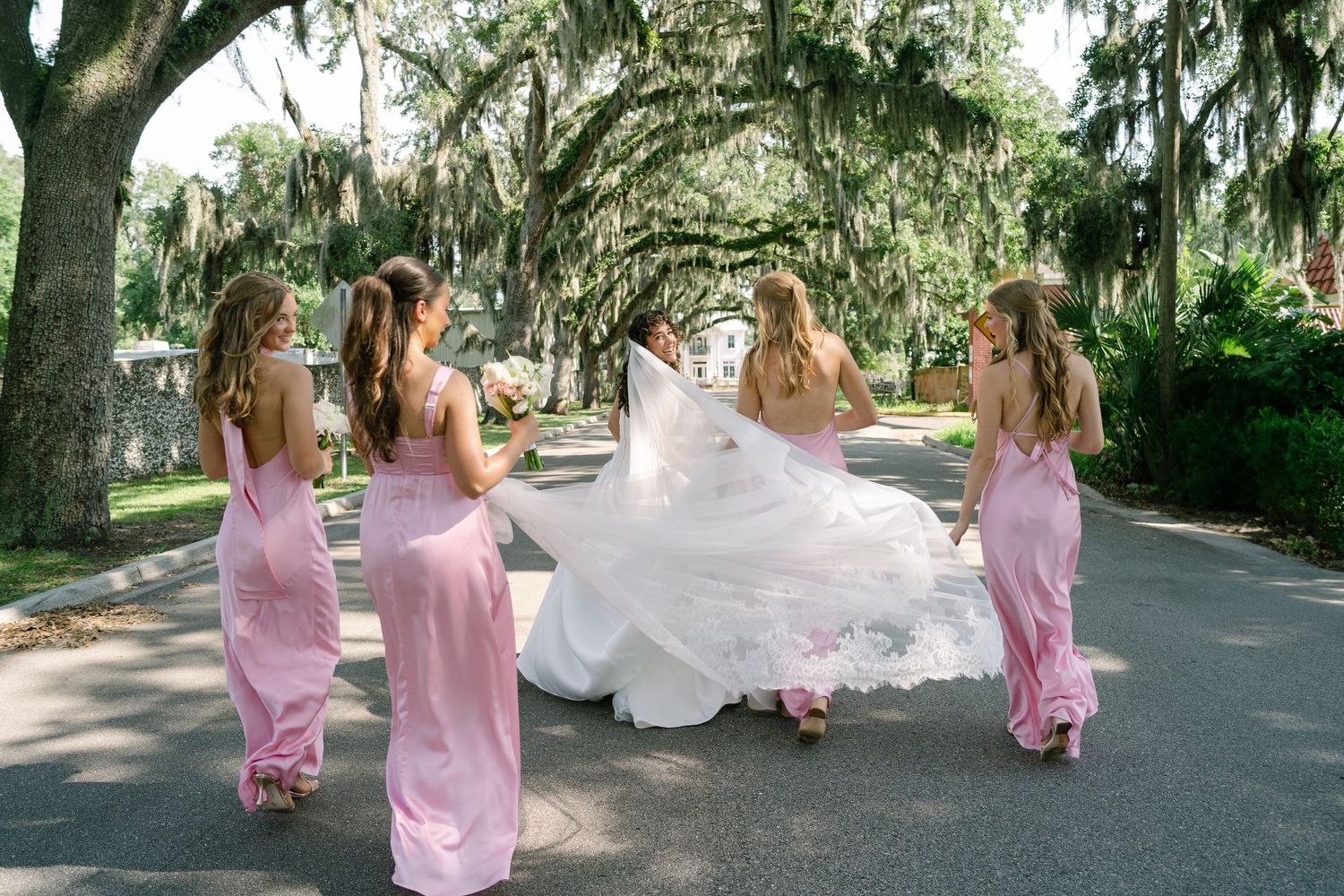 Bridesmaids in pink dresses playfully pose with bride's flowing dress on a moss-draped avenue.