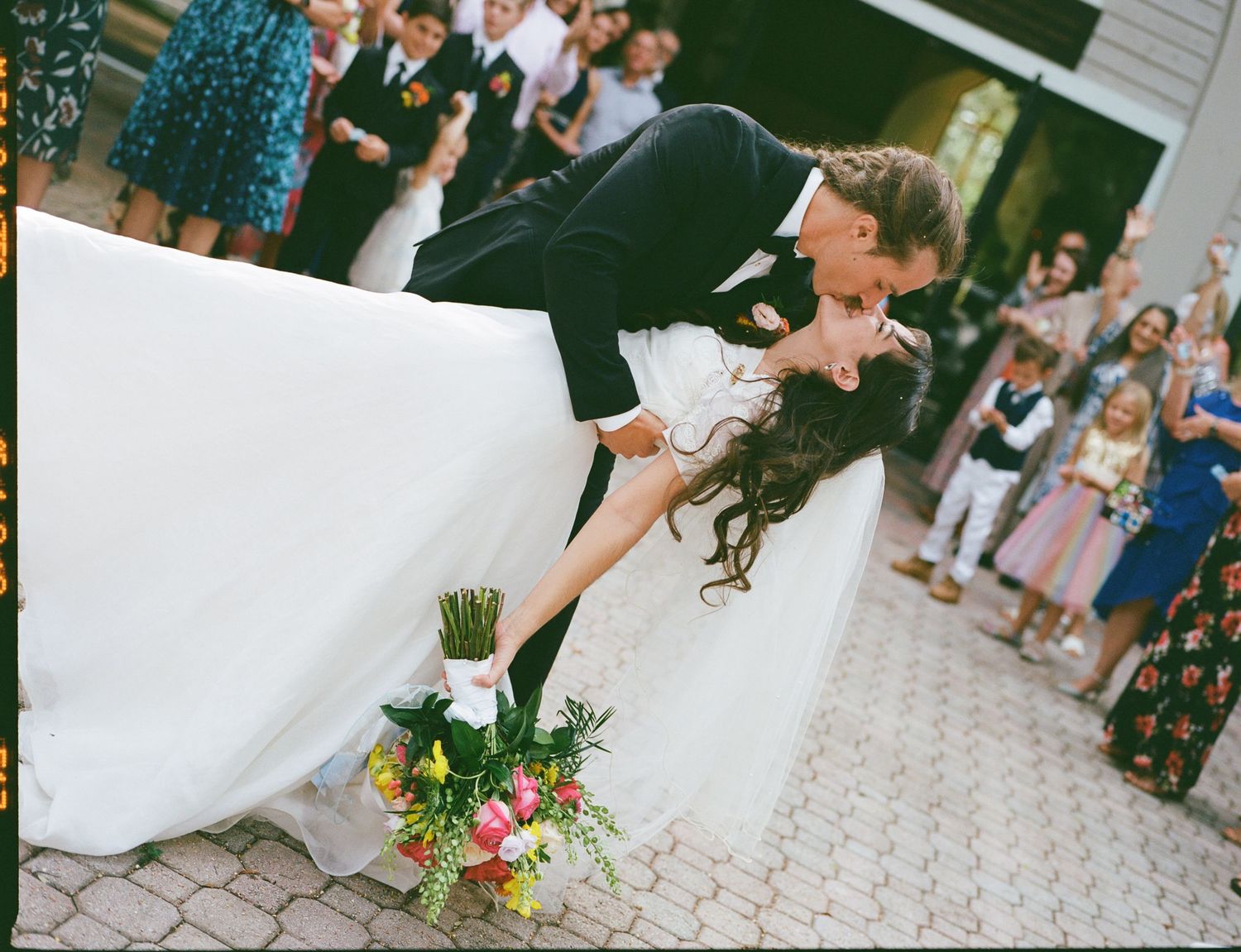 A dramatic dip kiss between newlyweds surrounded by wedding guests on a stone patio.