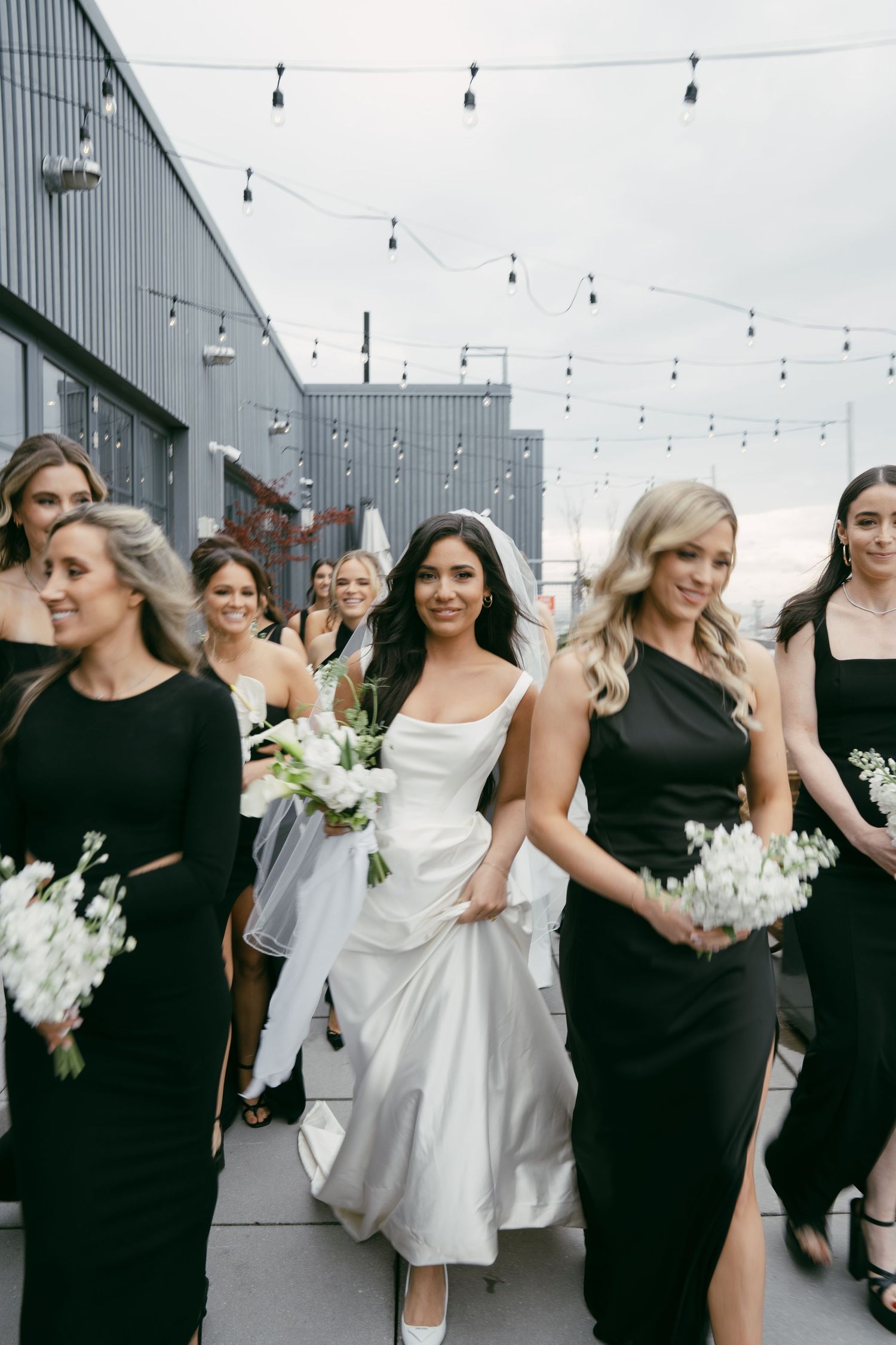 Bridal party walking in a line wearing black dresses and white bouquets under string lights outdoors.