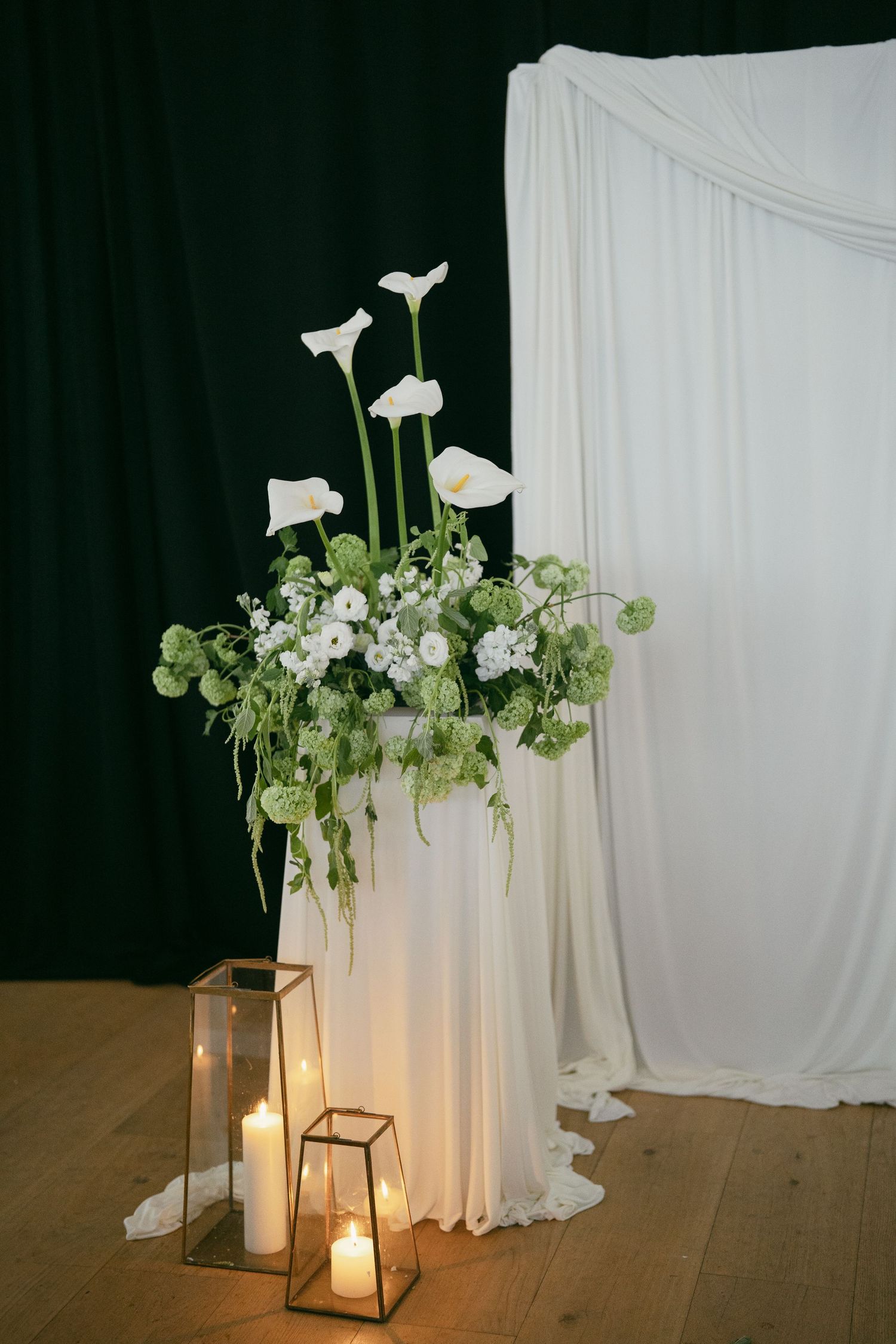 Elegant wedding decor featuring white calla lilies and greenery arrangements with illuminated lanterns against draped fabric.