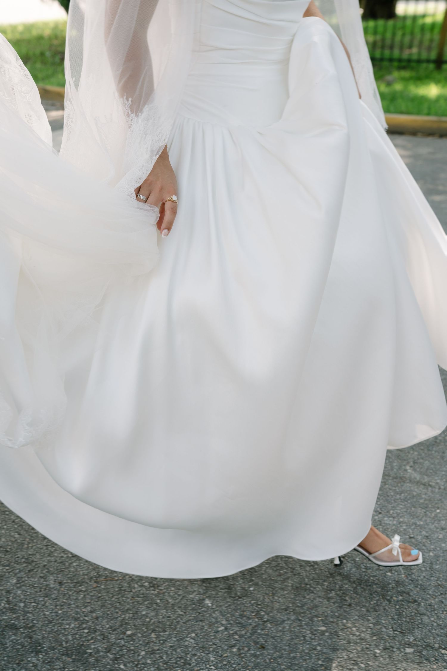 Close-up view of a flowing white wedding dress skirt and delicate sandals on a concrete surface.