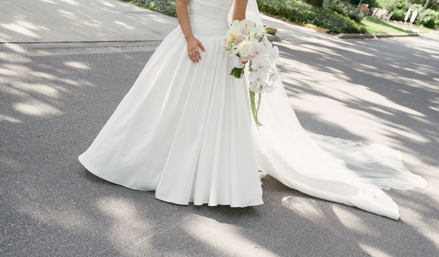 Flowing white wedding dress and bouquet detail photographed on a sun-dappled path.