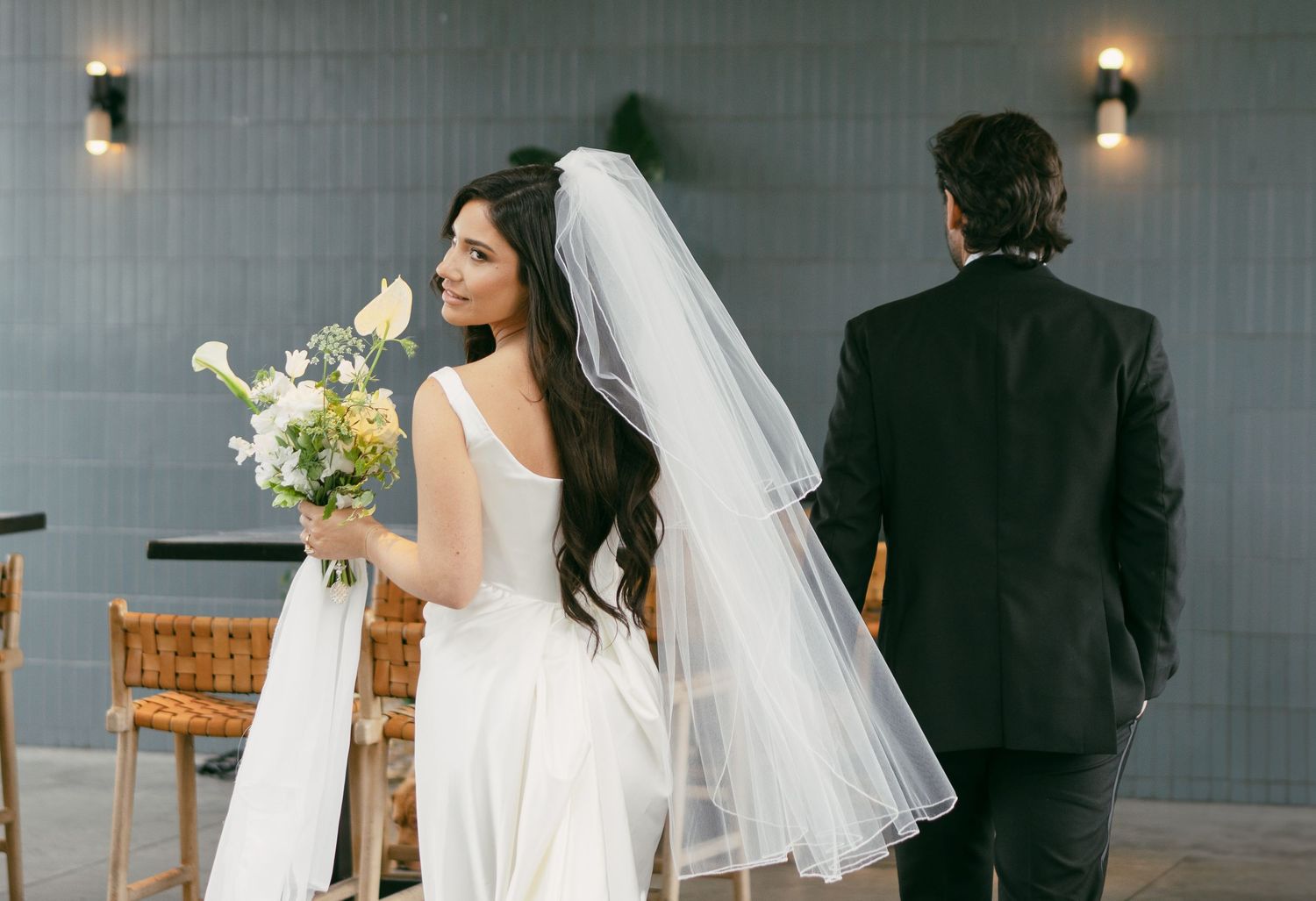 A bride in a sleek white dress holds a bouquet while standing in front of a grey wall.