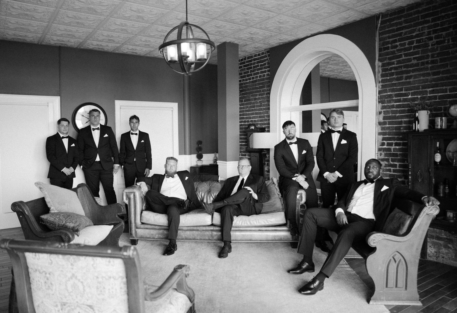 Groomsmen in black tuxedos lounging on vintage furniture for casual wedding party photo.
