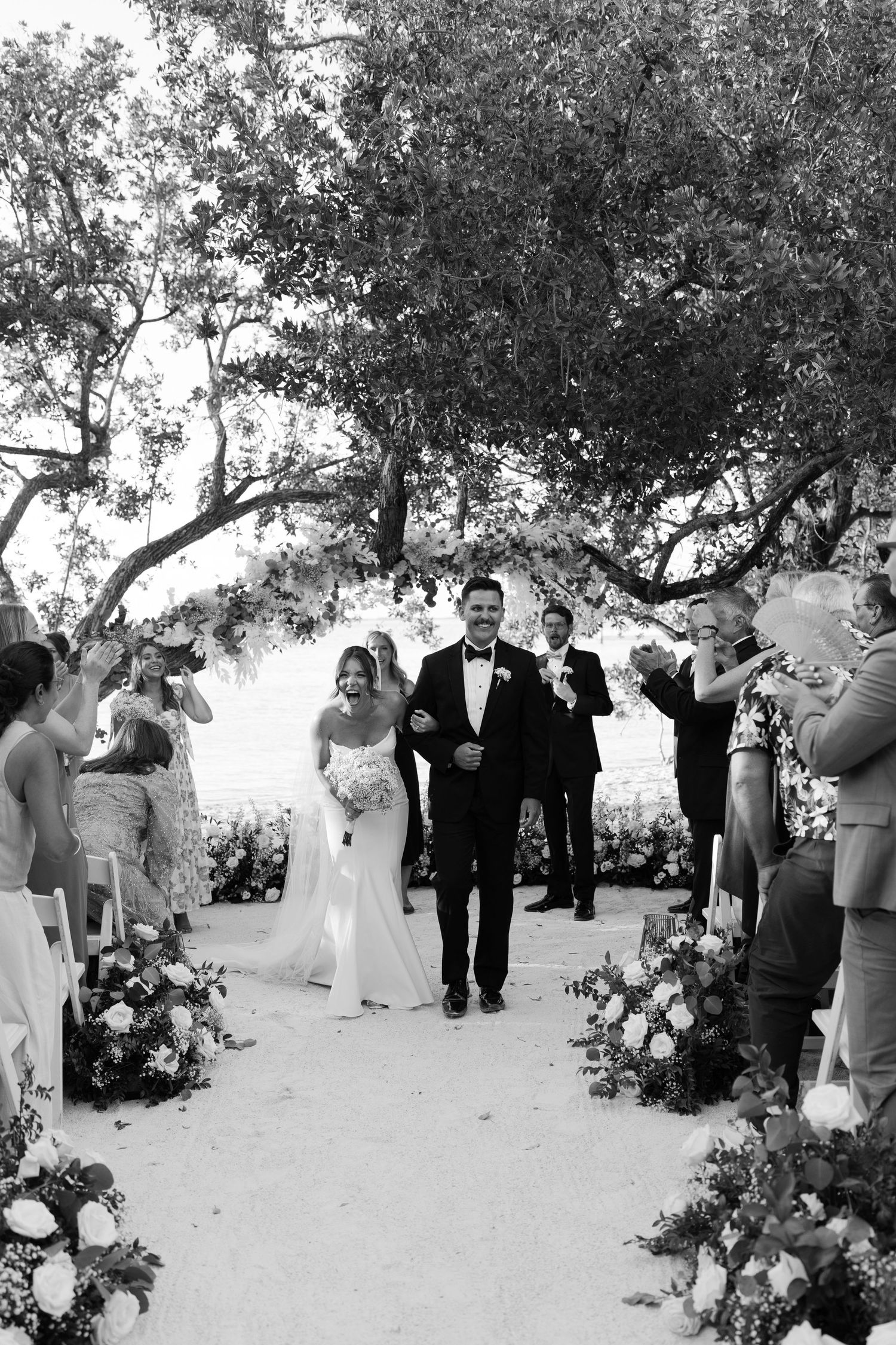 A couple walks down a flower-lined aisle during their outdoor wedding ceremony as guests look on.