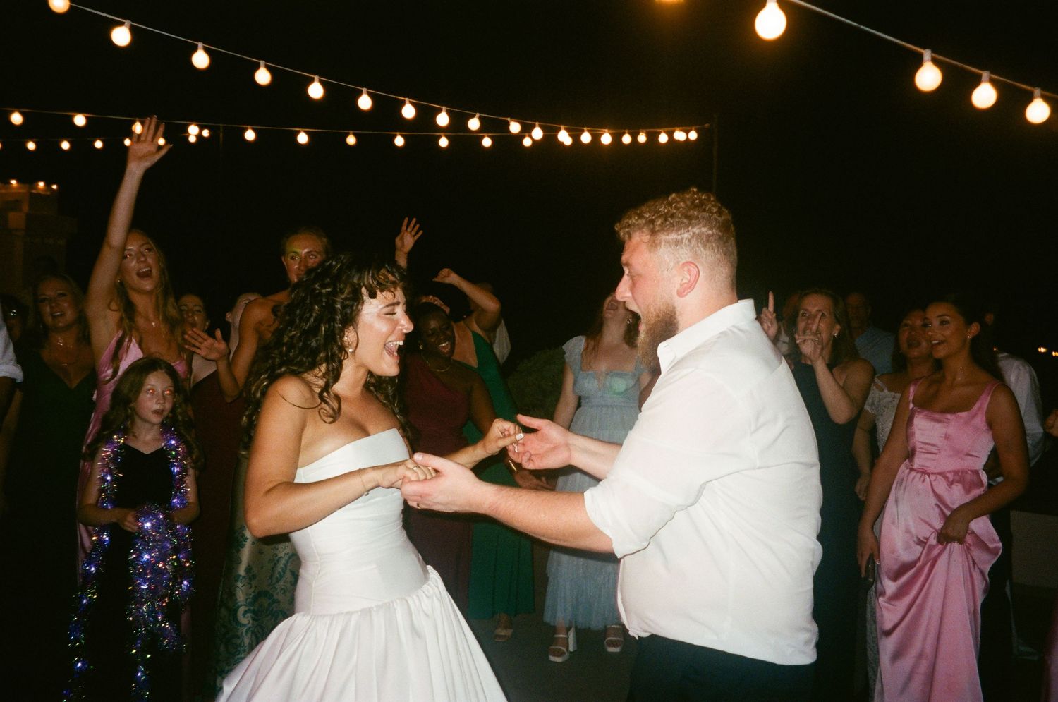 A couple dances energetically at their wedding reception under glowing string lights.