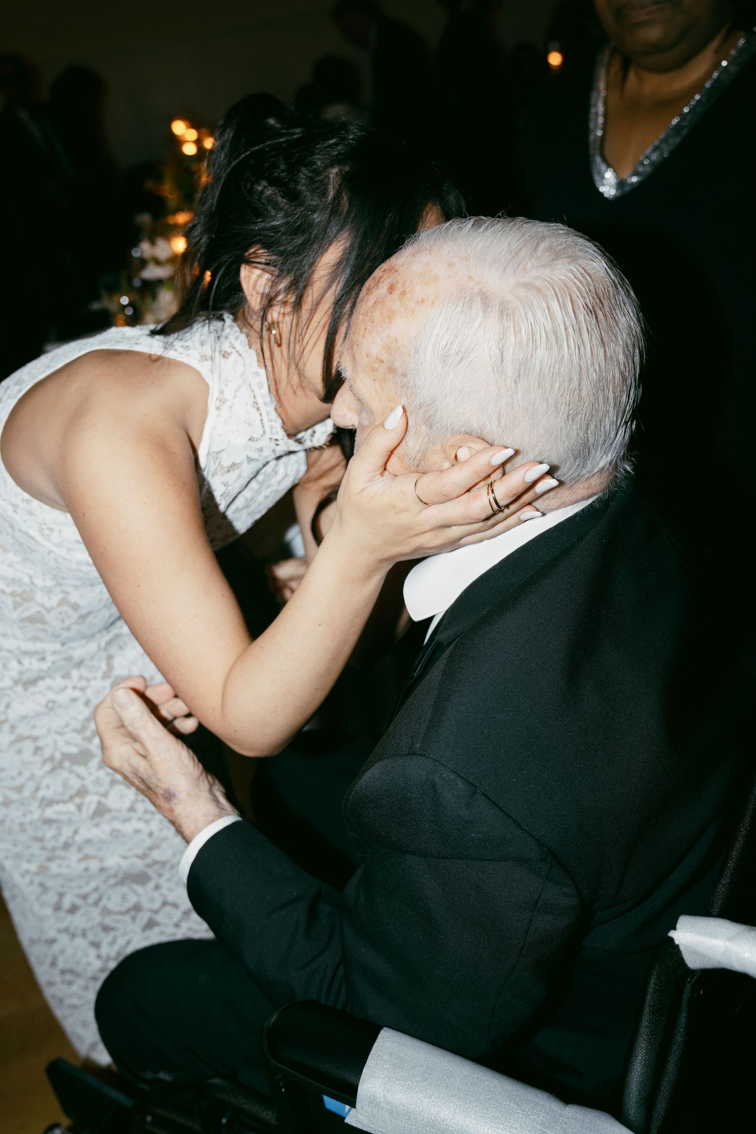 A tender moment shared between someone in a white wedding dress and an elderly guest in a wheelchair at a reception.