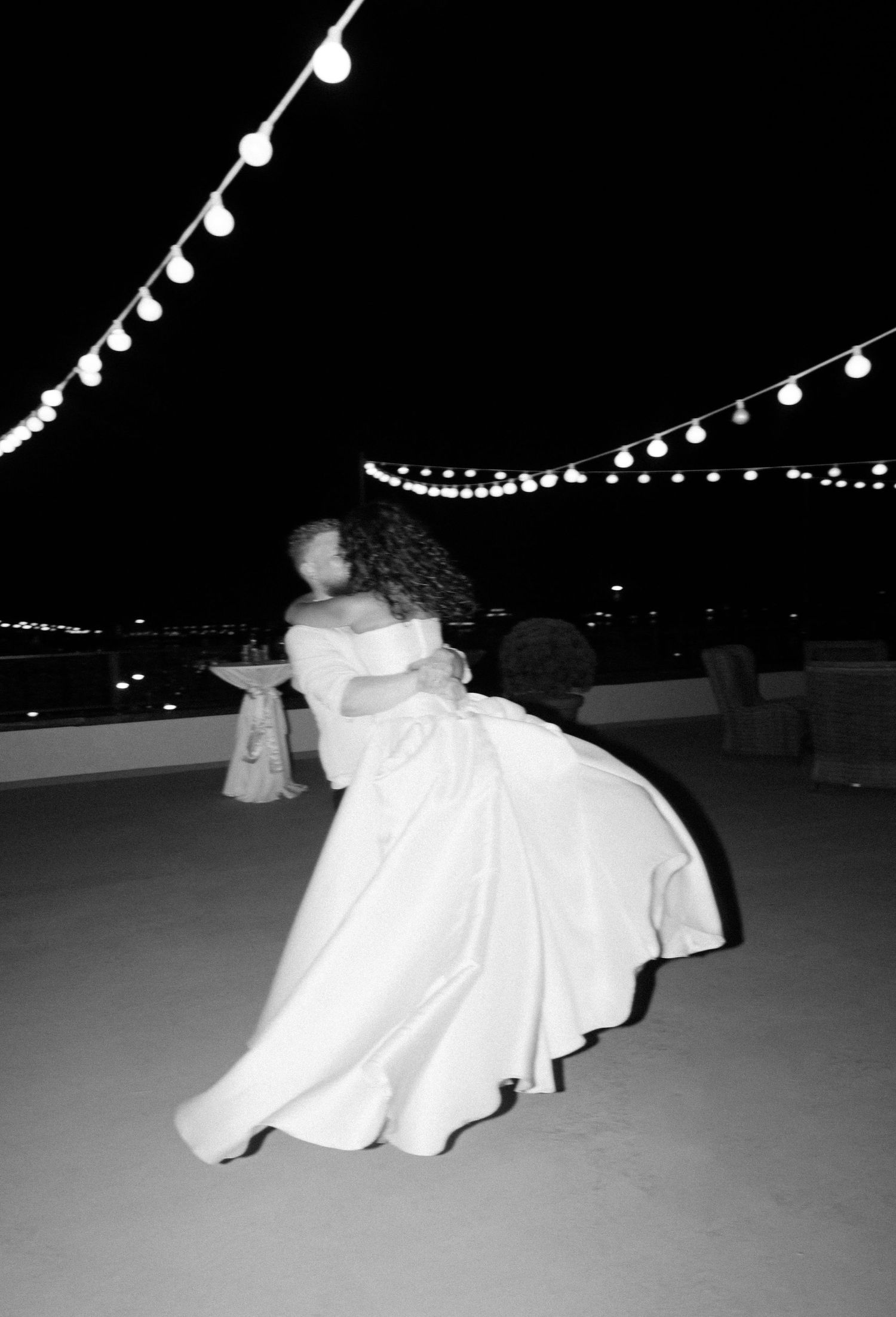 A romantic black and white dance photo under string lights at a rooftop wedding reception.
