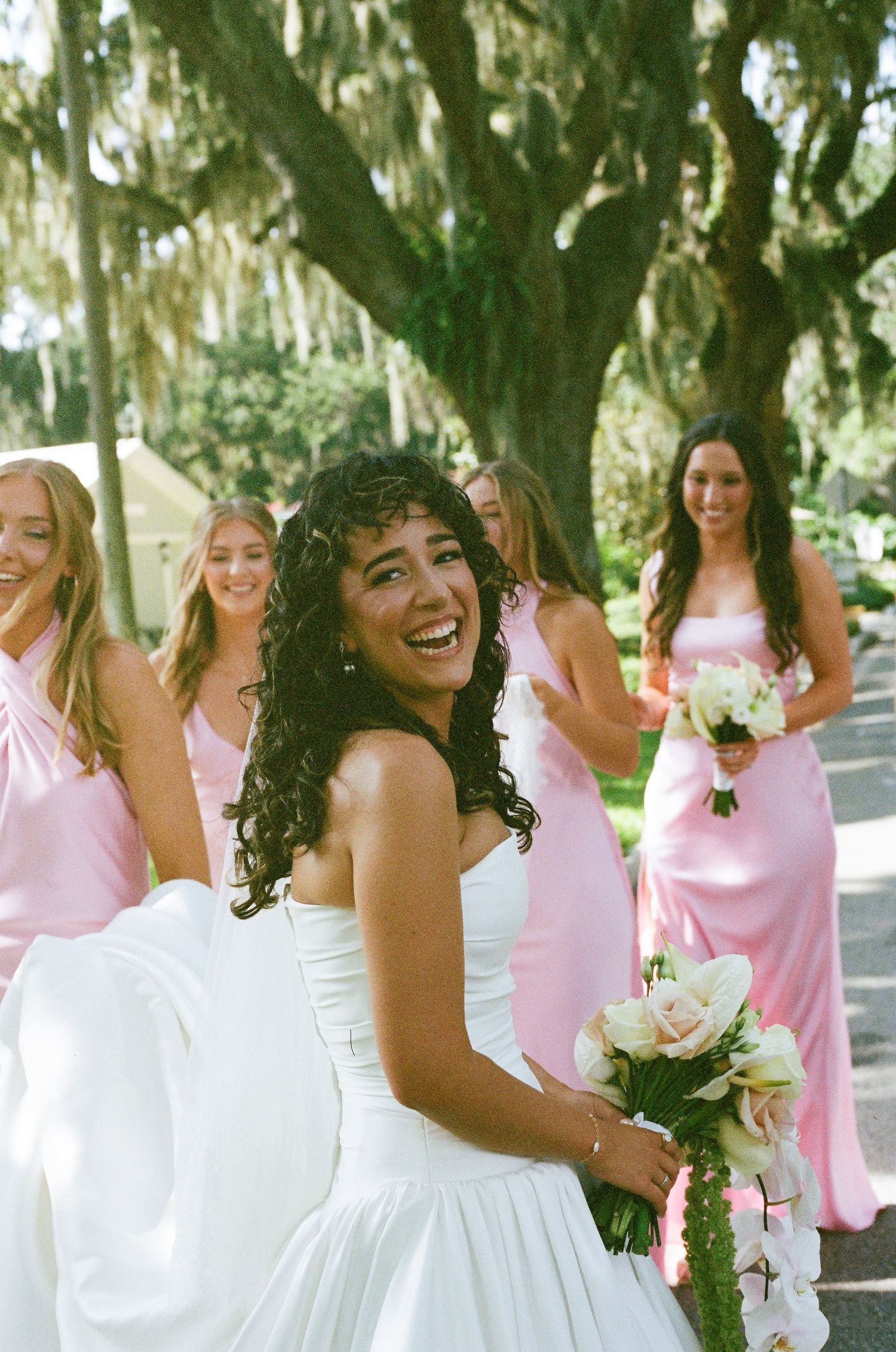 Wedding party poses joyfully under oak trees with bridesmaids in pink dresses.