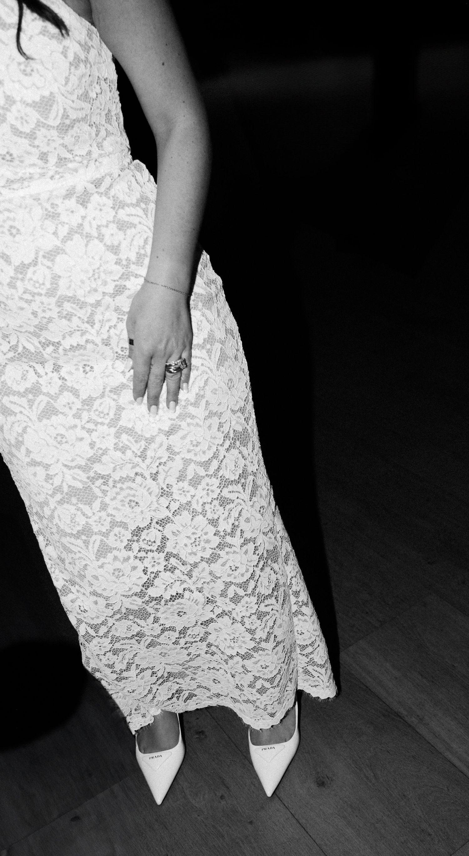 A person wearing a white lace dress and pointed heels stands on a dark wooden floor.