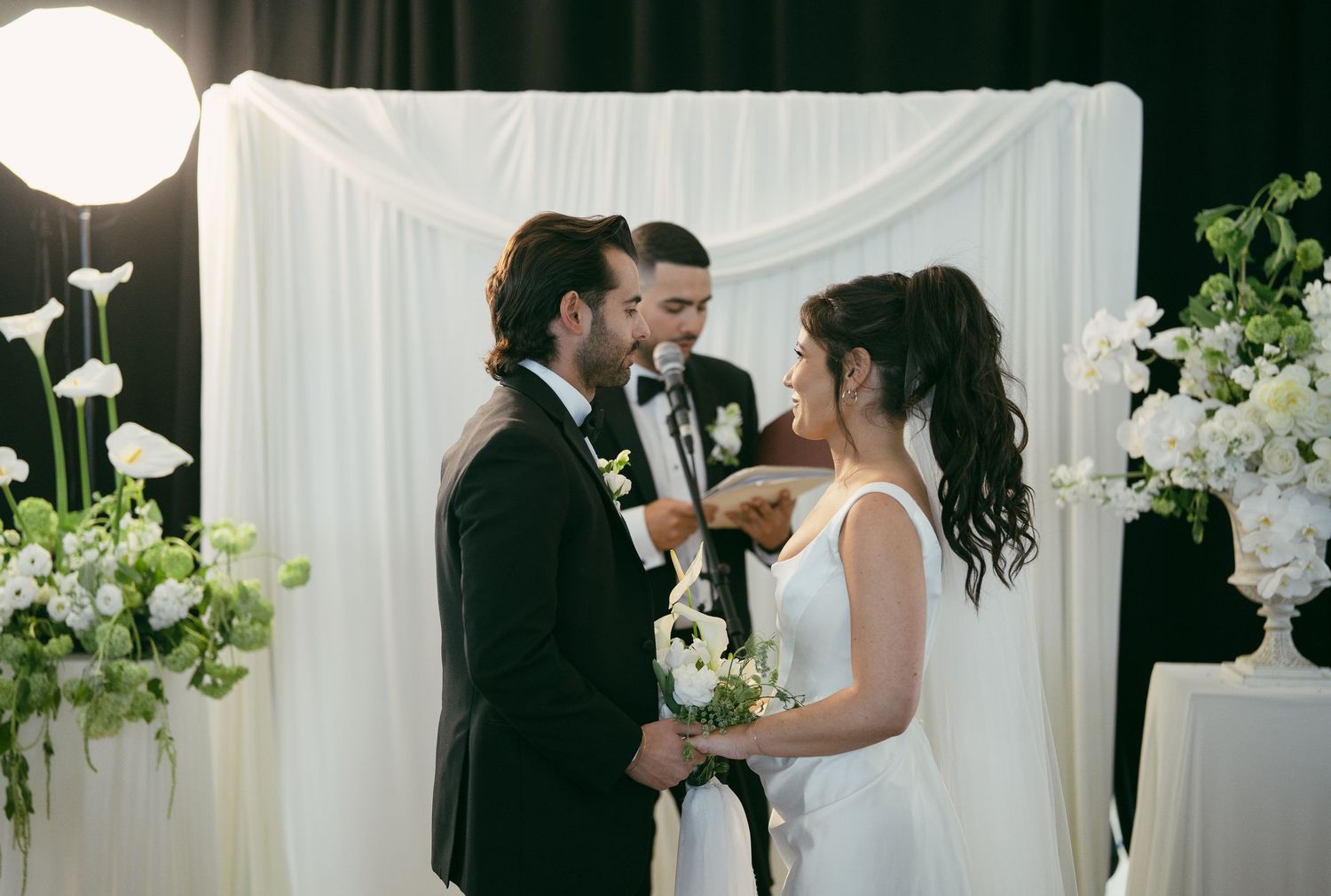 Wedding ceremony setup with white floral arrangements and drapery during an indoor celebration.