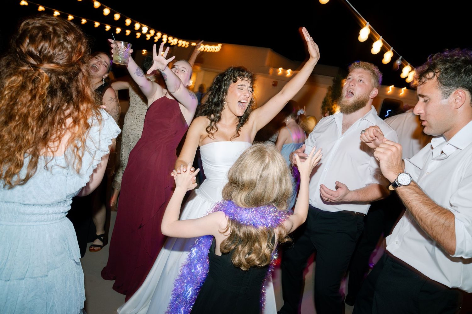Wedding reception guests dancing and celebrating under warm string lights at night.