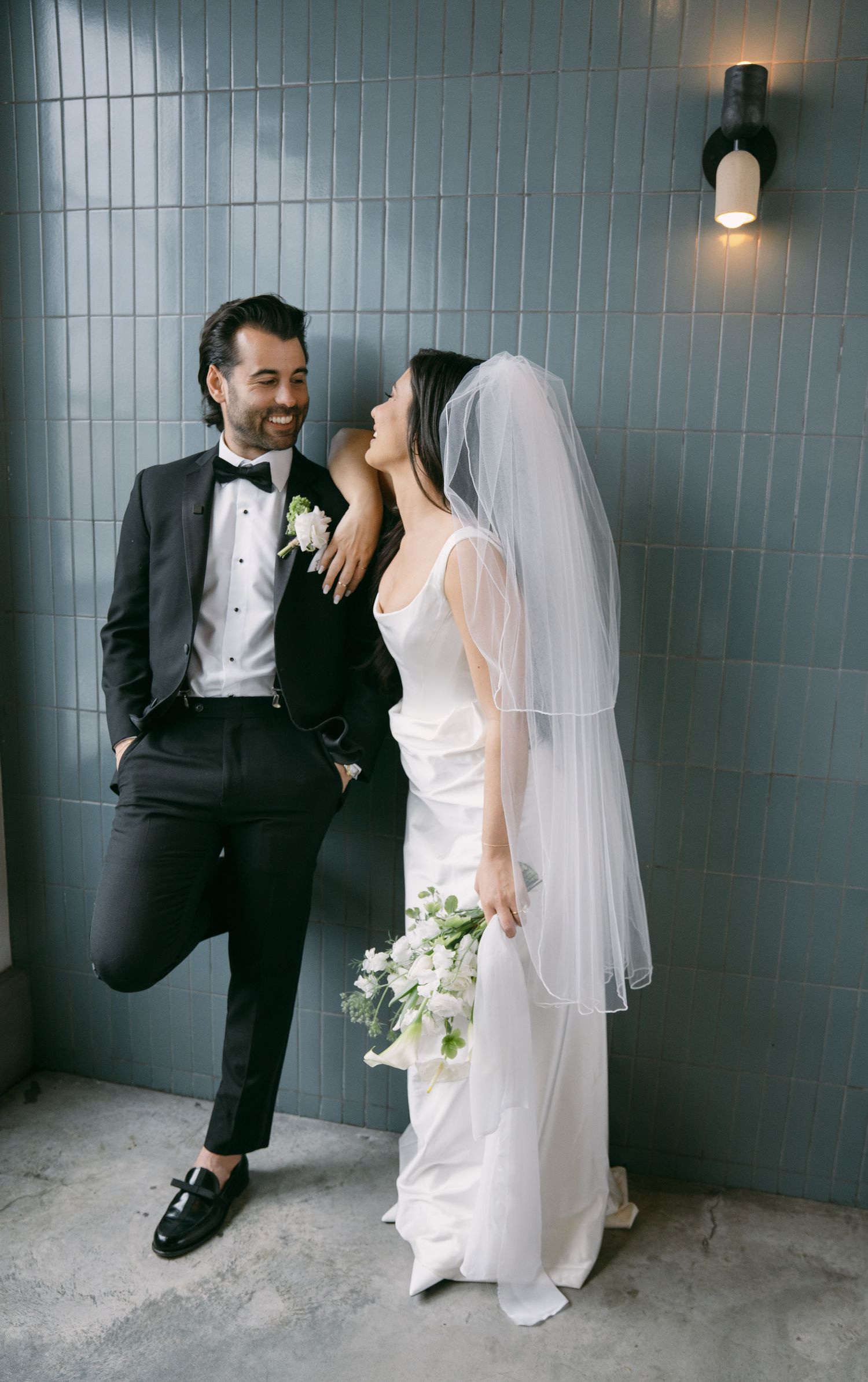 A newlywed couple share a candid moment against a grey tiled wall, with the bride holding a white bouquet.