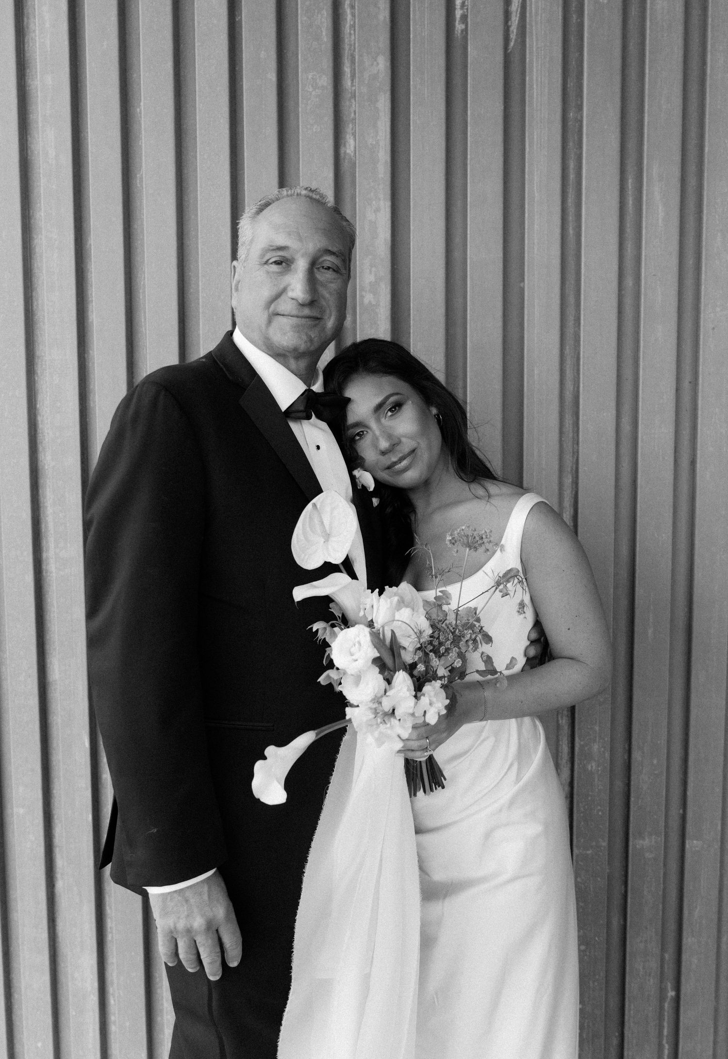 Black and white wedding portrait of a couple in formal attire standing against a striped wall.