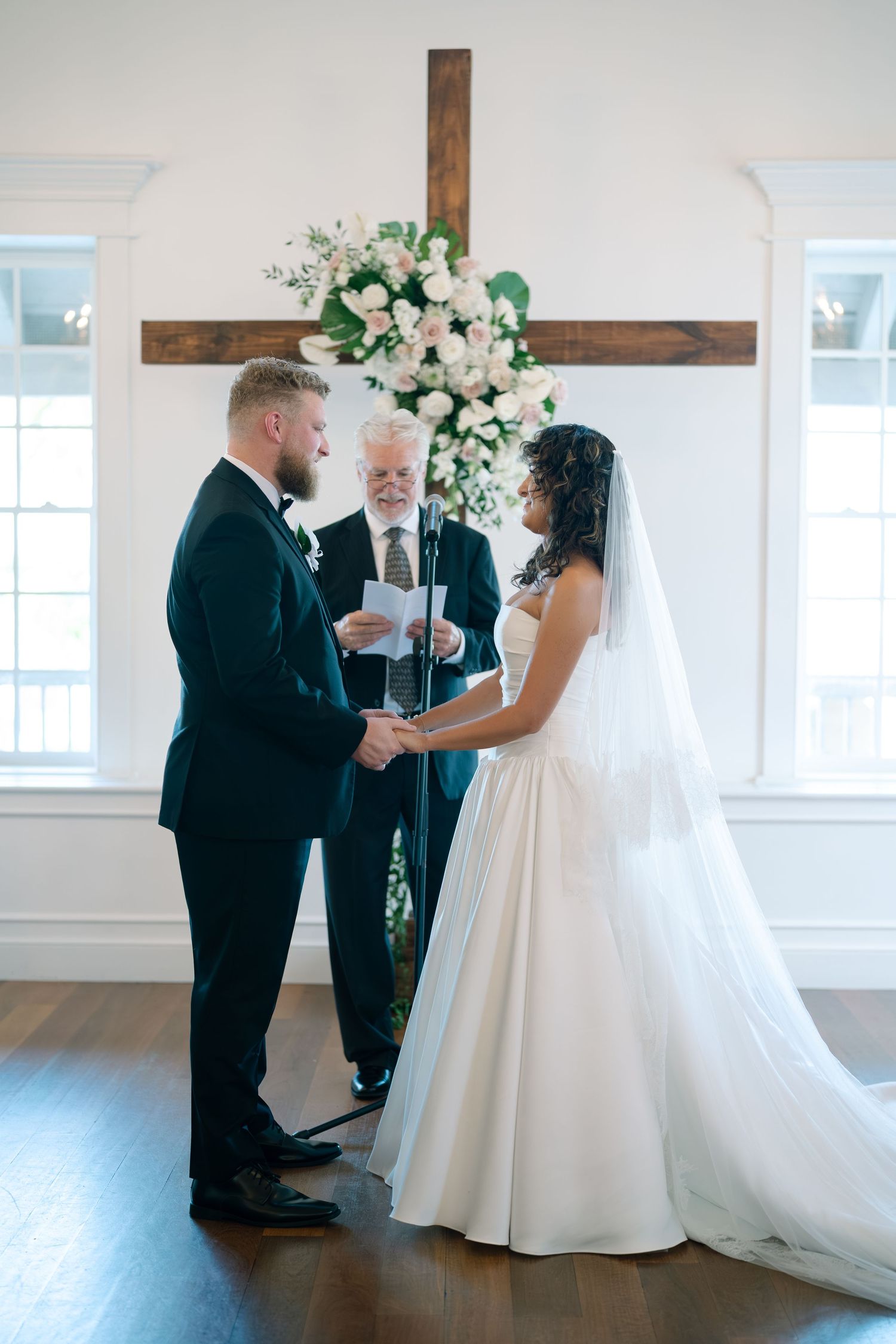 Wedding ceremony in a bright chapel with wooden cross and floral arrangement decorating the altar.
