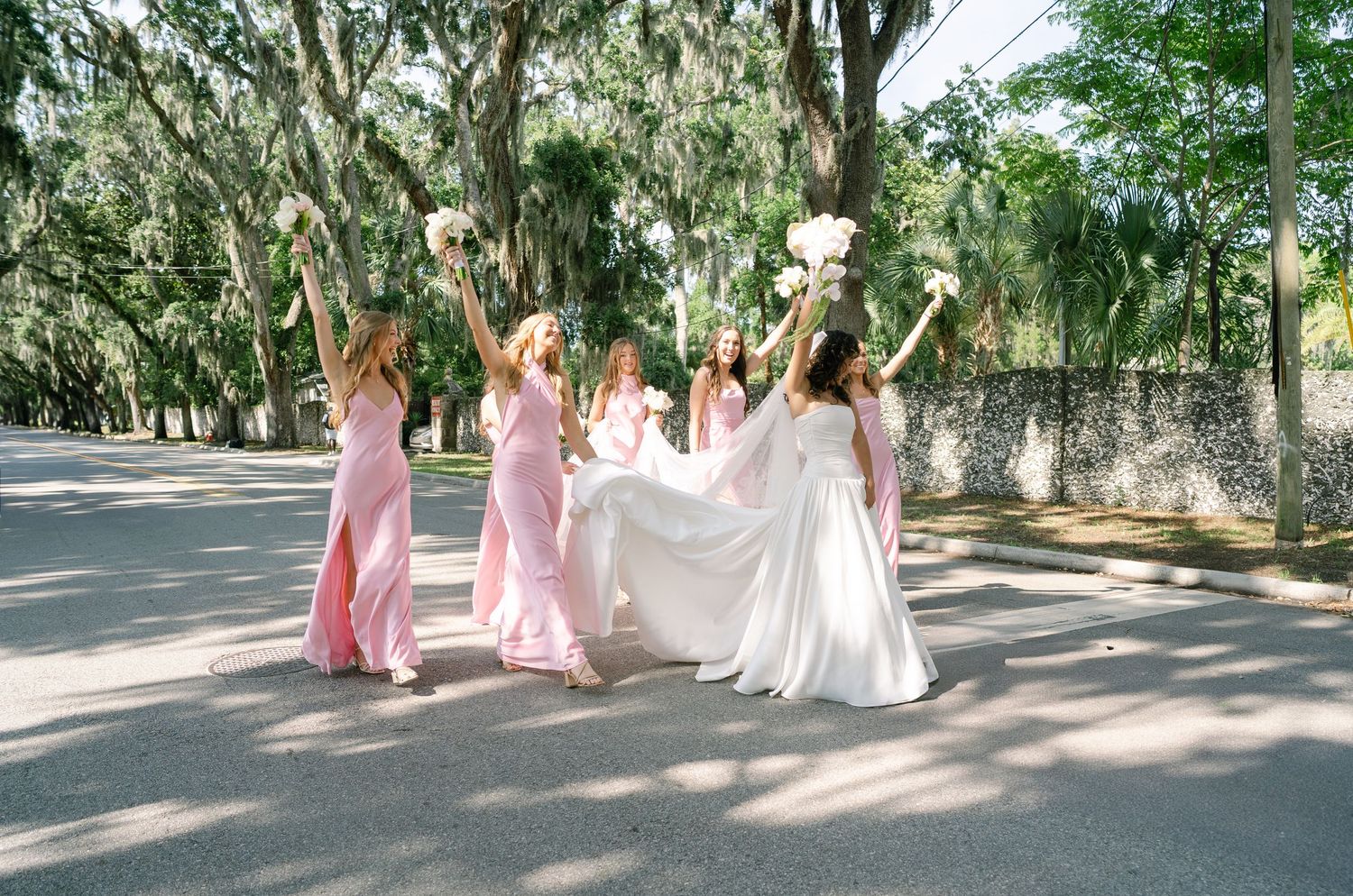 Bridesmaids in pink gowns help lift the bride's dress while walking under moss-draped trees.
