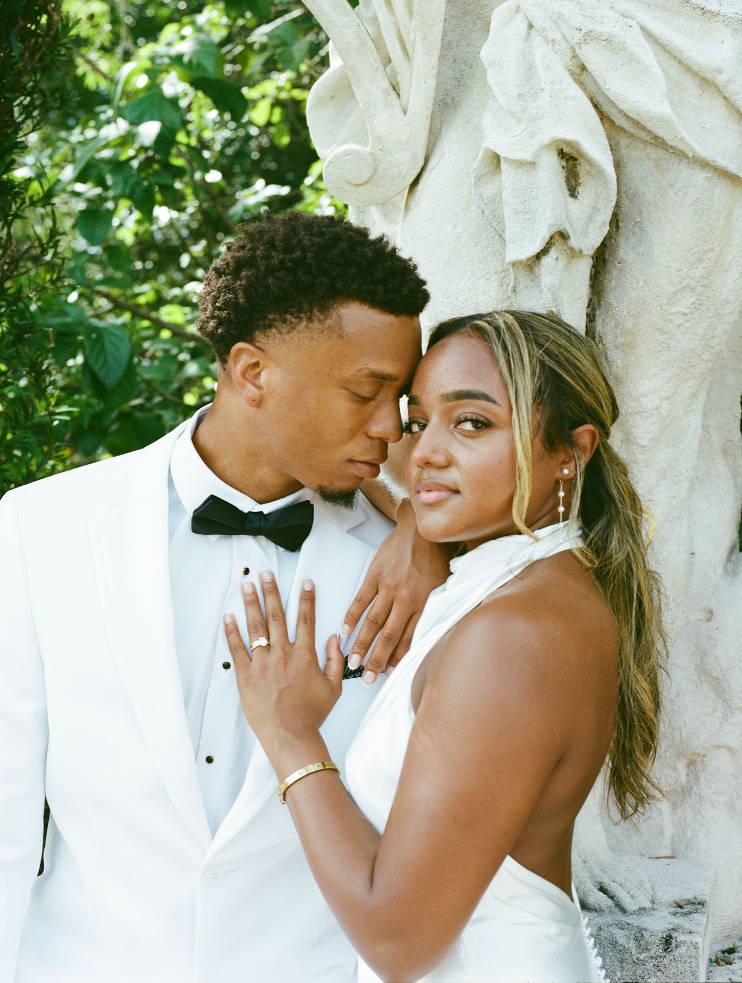 A couple in formal white wedding attire share an intimate moment outdoors by an ornate stone column.