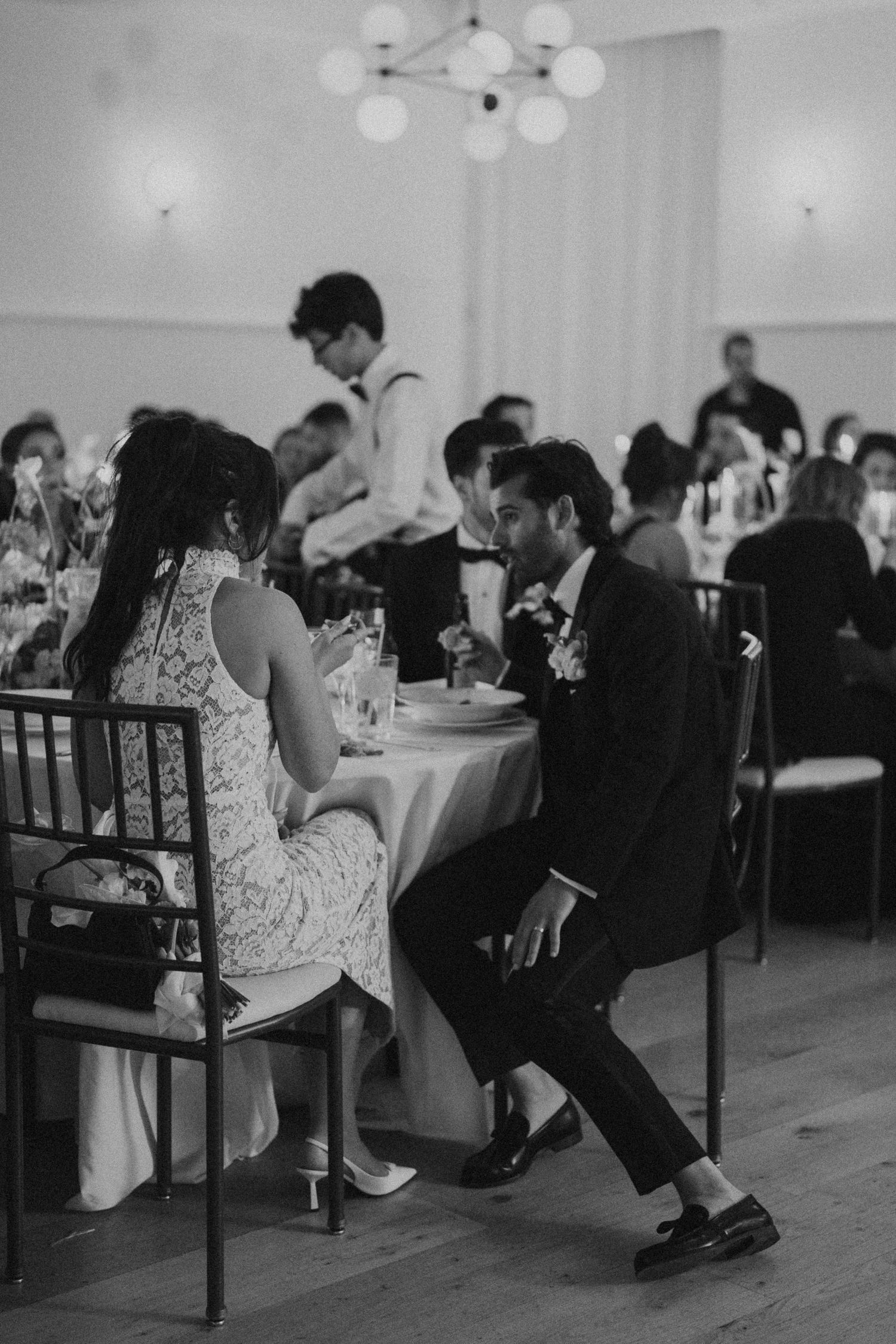 Wedding guests seated at dinner table in a dimly lit reception hall with overhead chandelier.