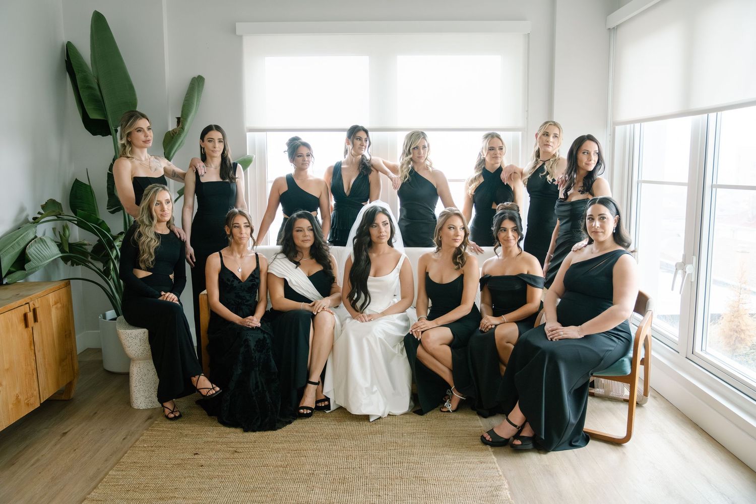 A group in coordinated black dresses and one in white sit together in a bright sunlit room with large windows and plants.